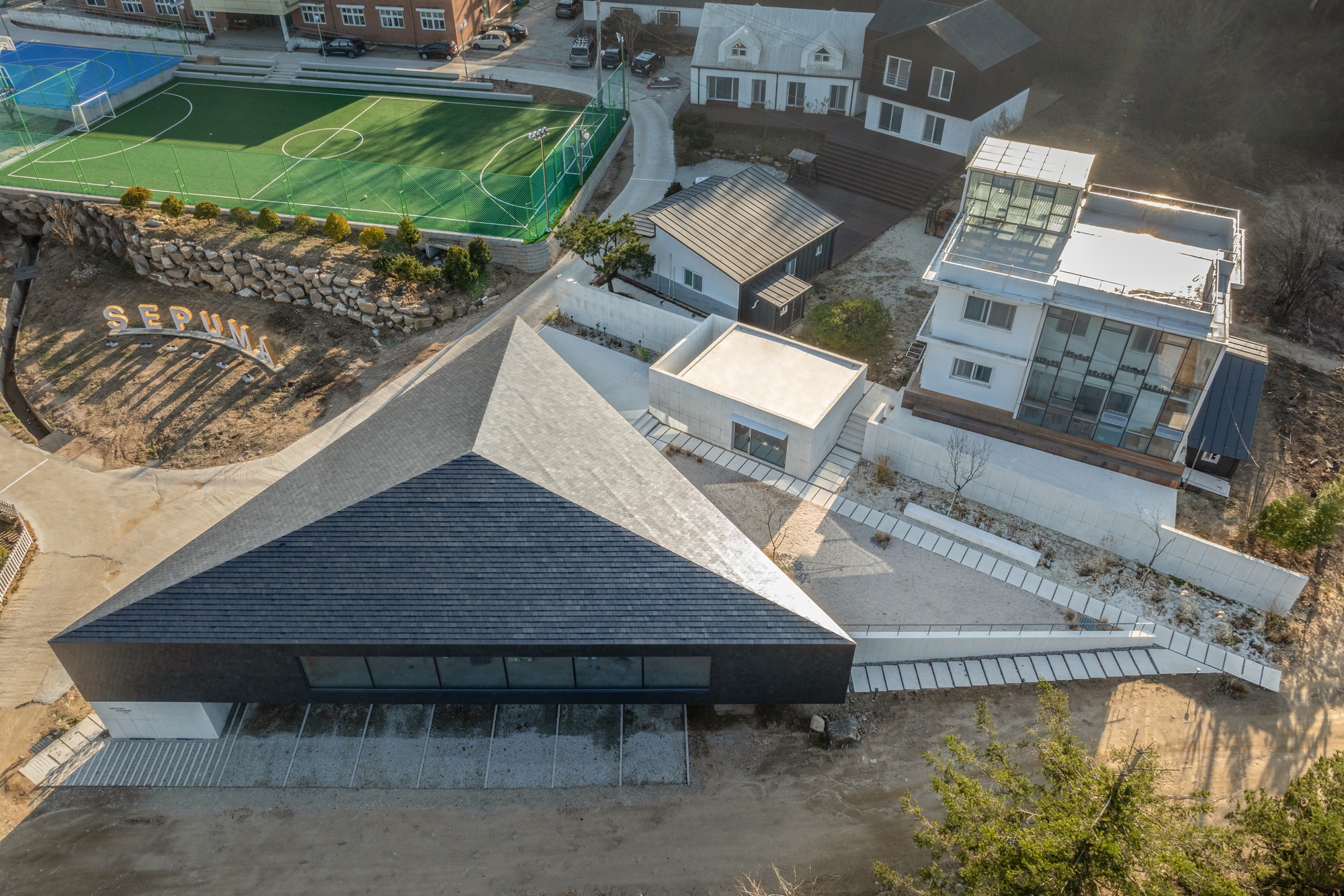 Aerial view of pyramidal slate roof and adjoining white volumes next to a sports field