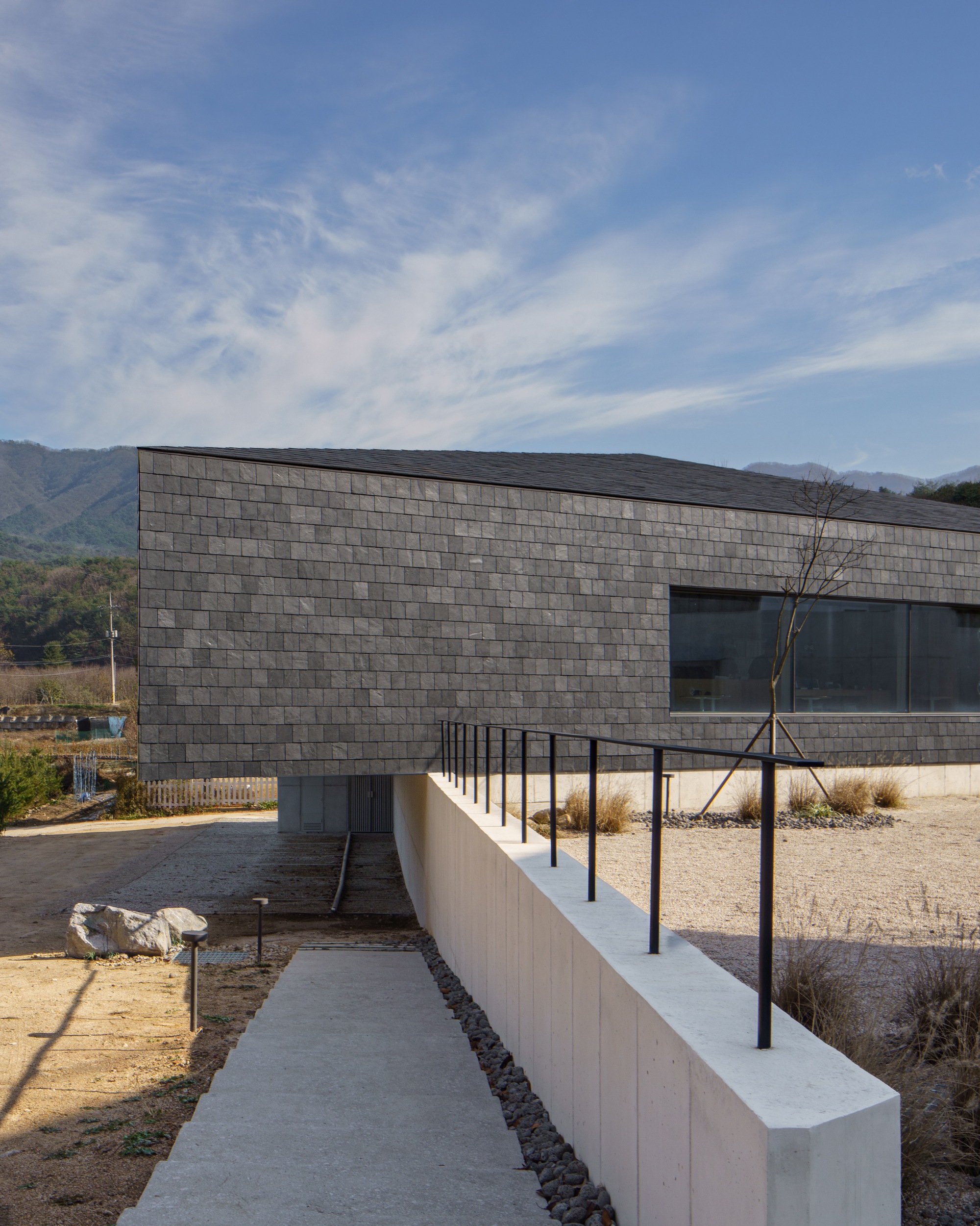 Concrete path leading to a dark stone tile facade with white retaining wall and steel railings