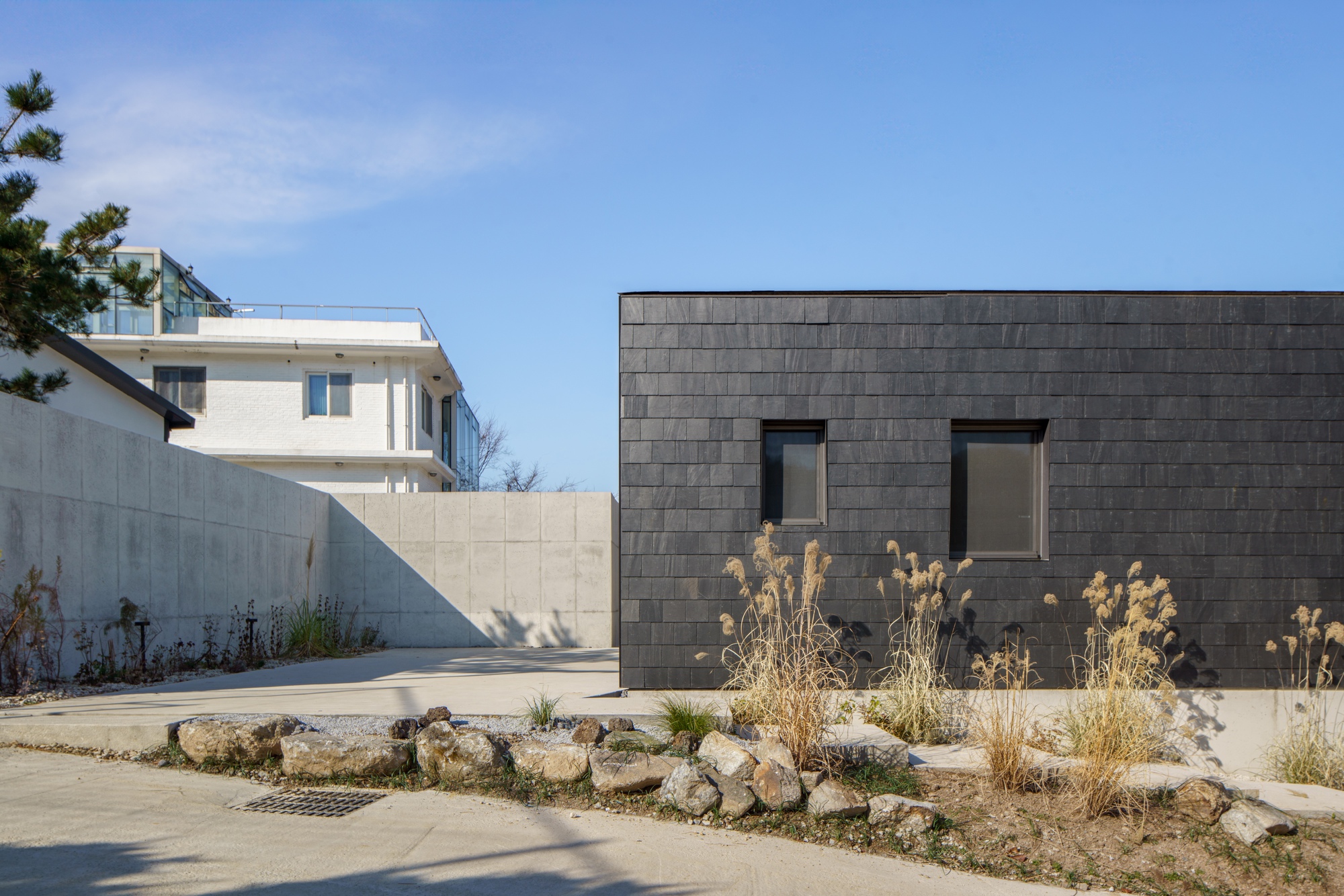 Courtyard view of dark stone tile volume with two small windows and winter grasses in foreground