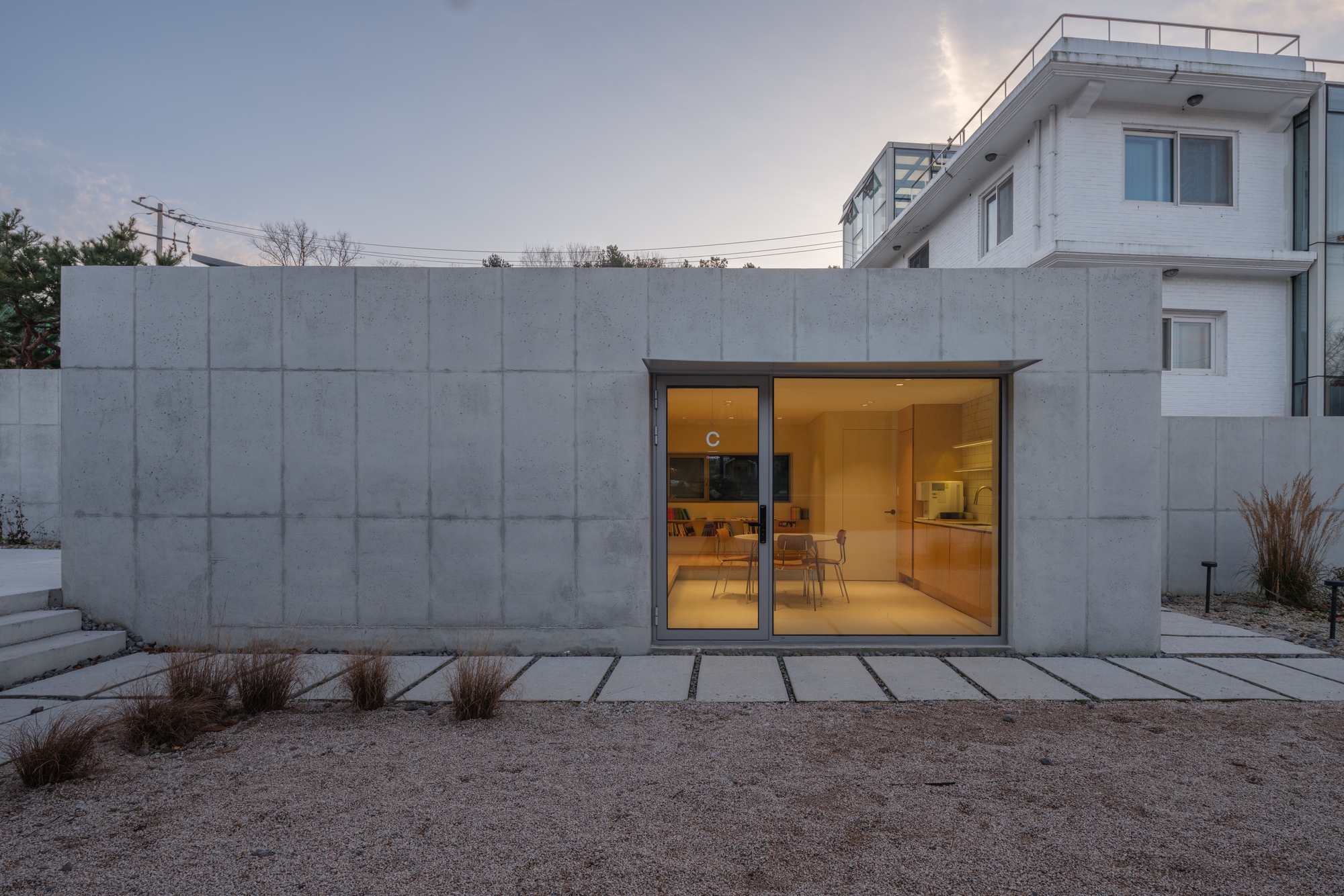 Concrete panel facade with glazed entry opening and paver pathway under a pale evening sky