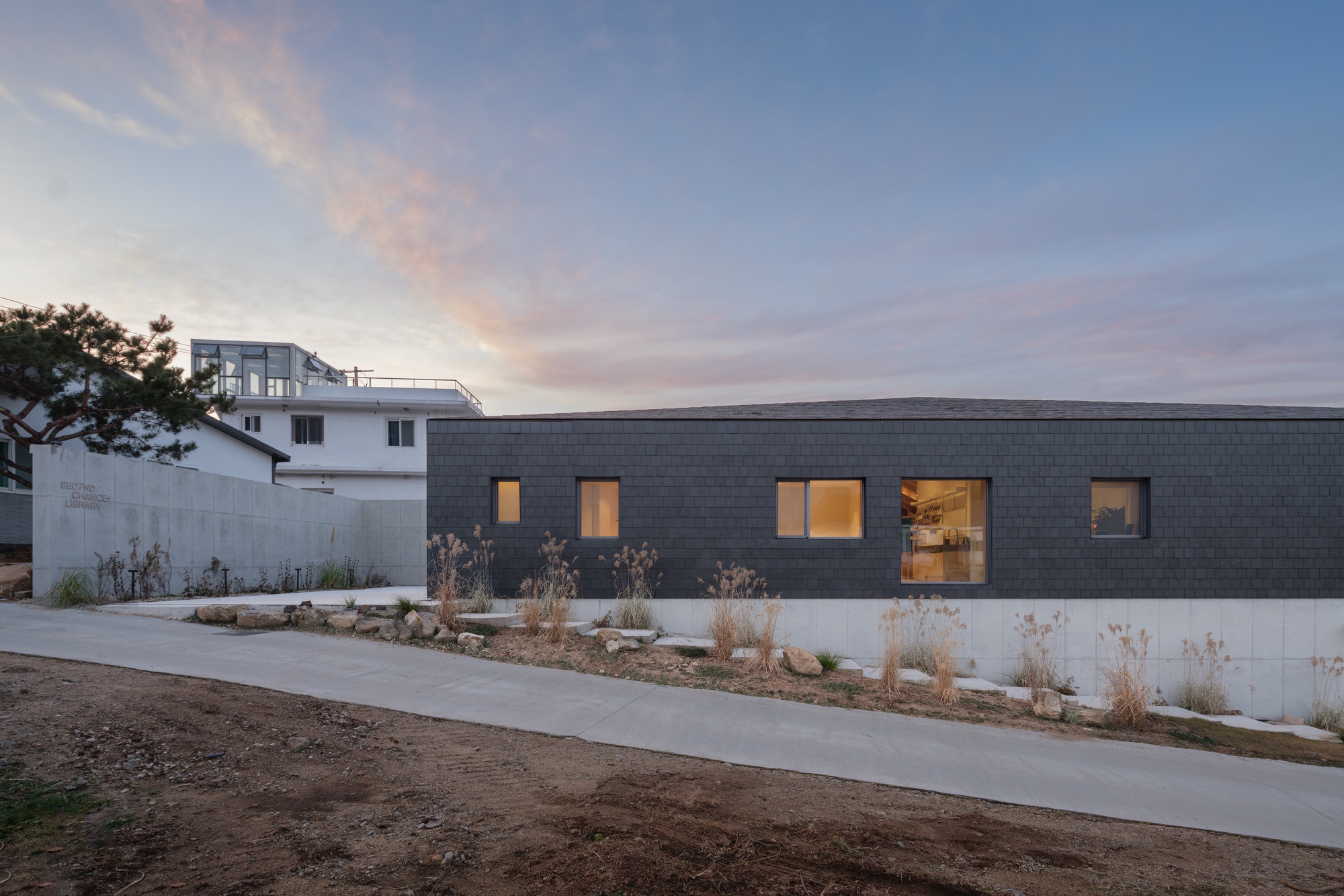 Dark tile-clad facade with punched windows and planted bed at dusk