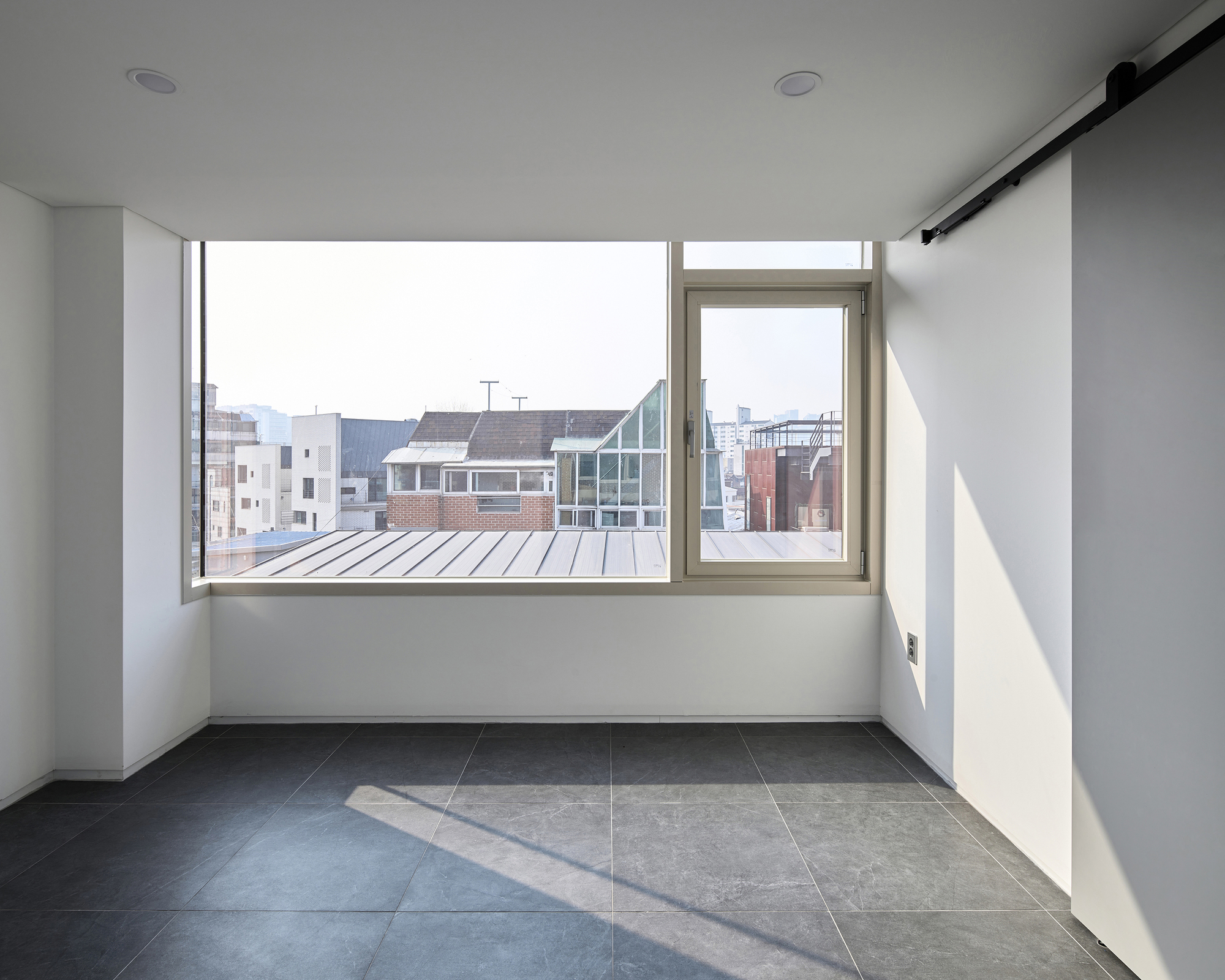 Empty room with large corner windows framing views of neighboring brick and glass buildings in daylight