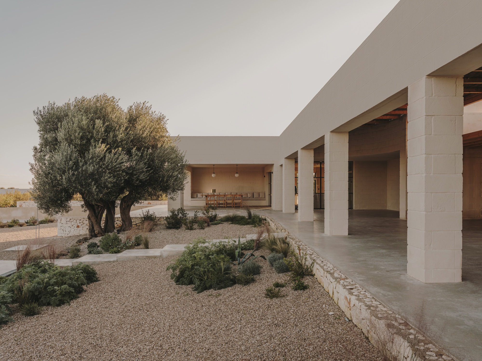 Courtyard with gravel and planted beds beside a covered colonnade at dusk