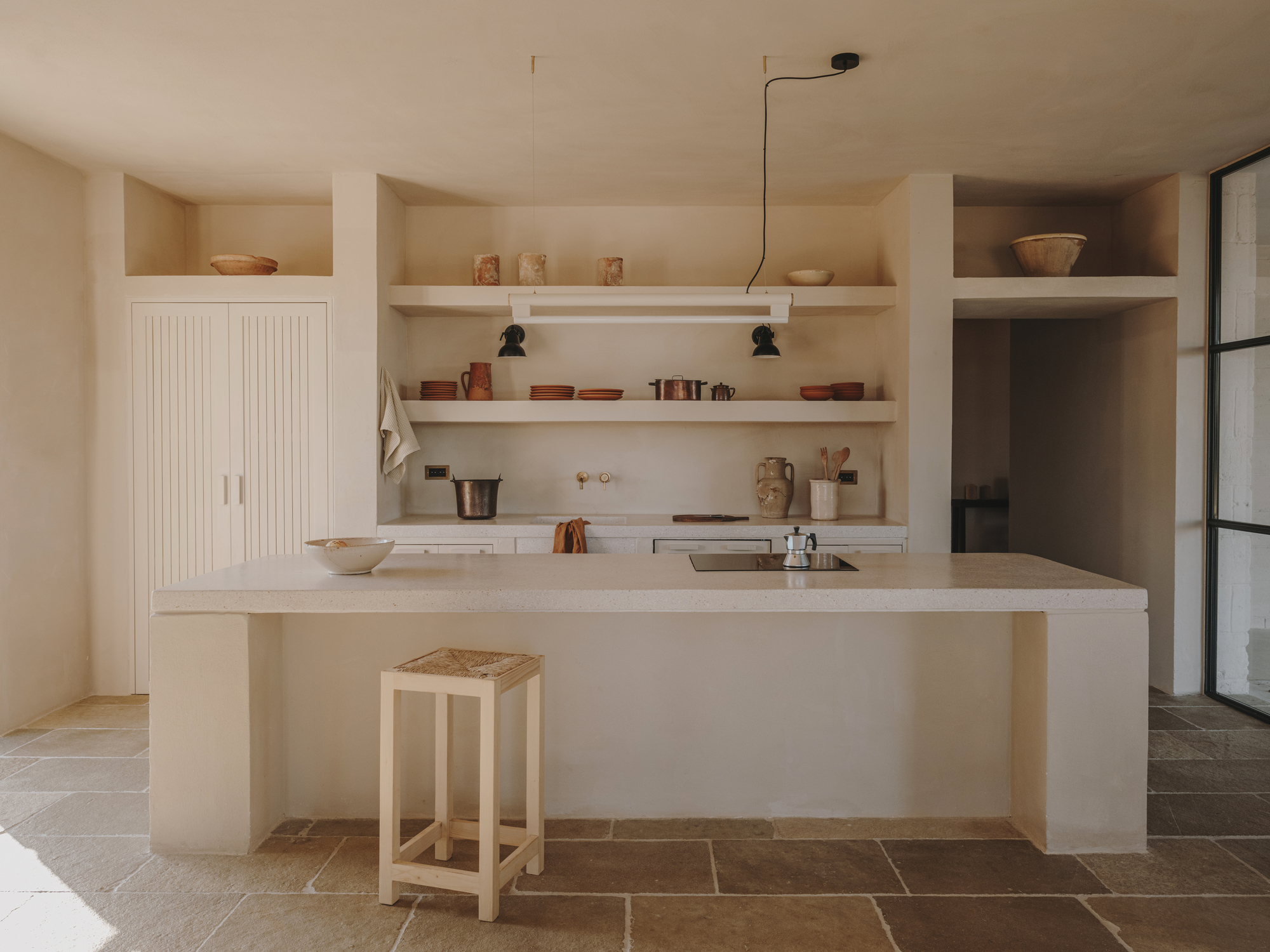 Kitchen with pale plaster island, open shelving and grooved cabinet doors on terracotta tile floor