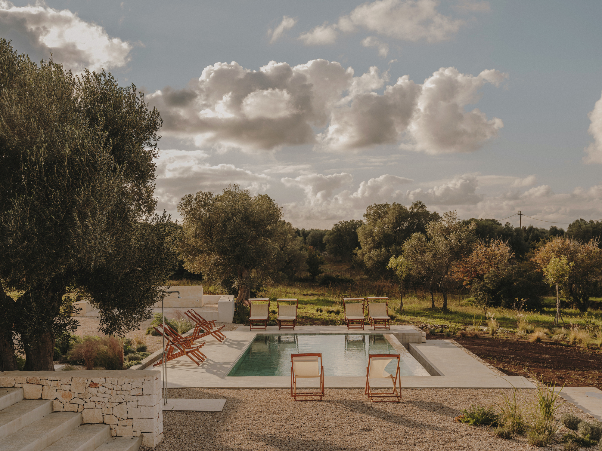Pool terrace edged by dry stone walls with olive groves beyond under dramatic sky