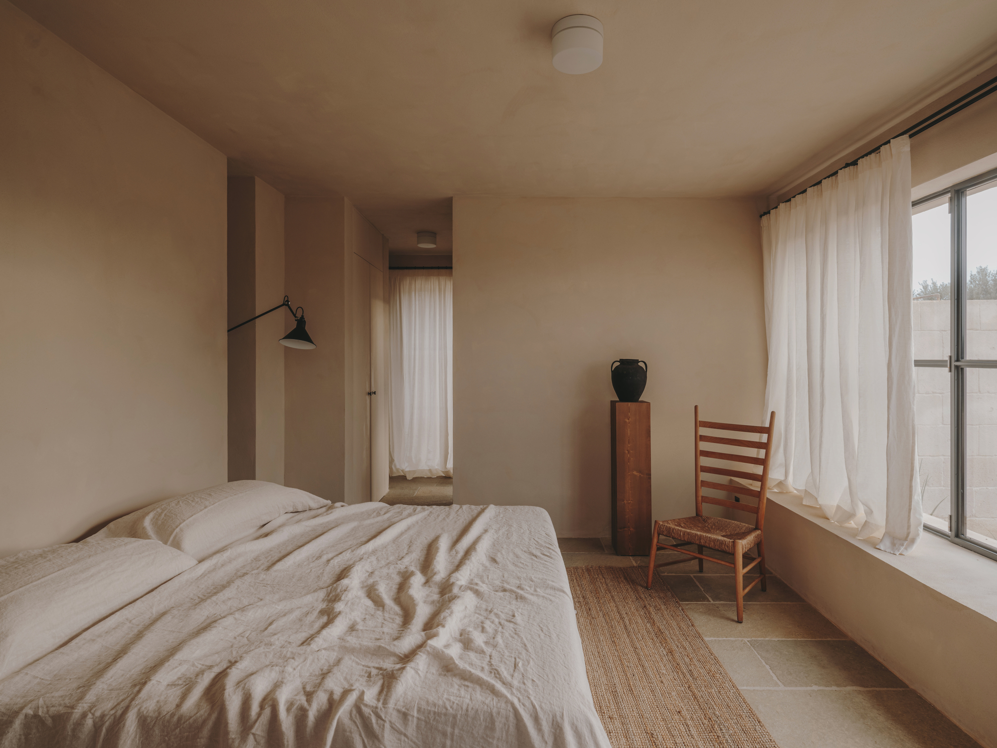 Bedroom with pale plaster walls, sheer curtains and a ladder-back chair beside a timber cabinet