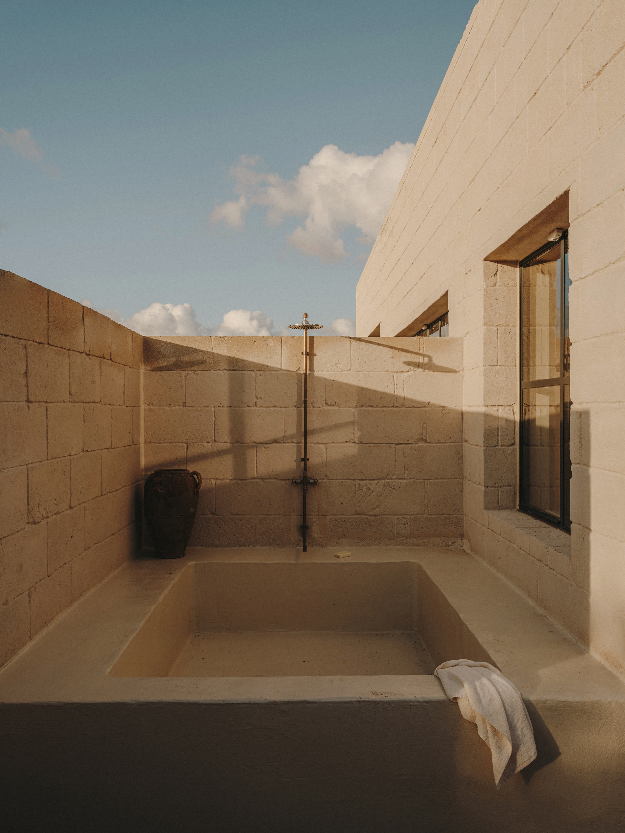 Outdoor sunken bathtub enclosed by concrete block walls with exposed brass plumbing under afternoon sky