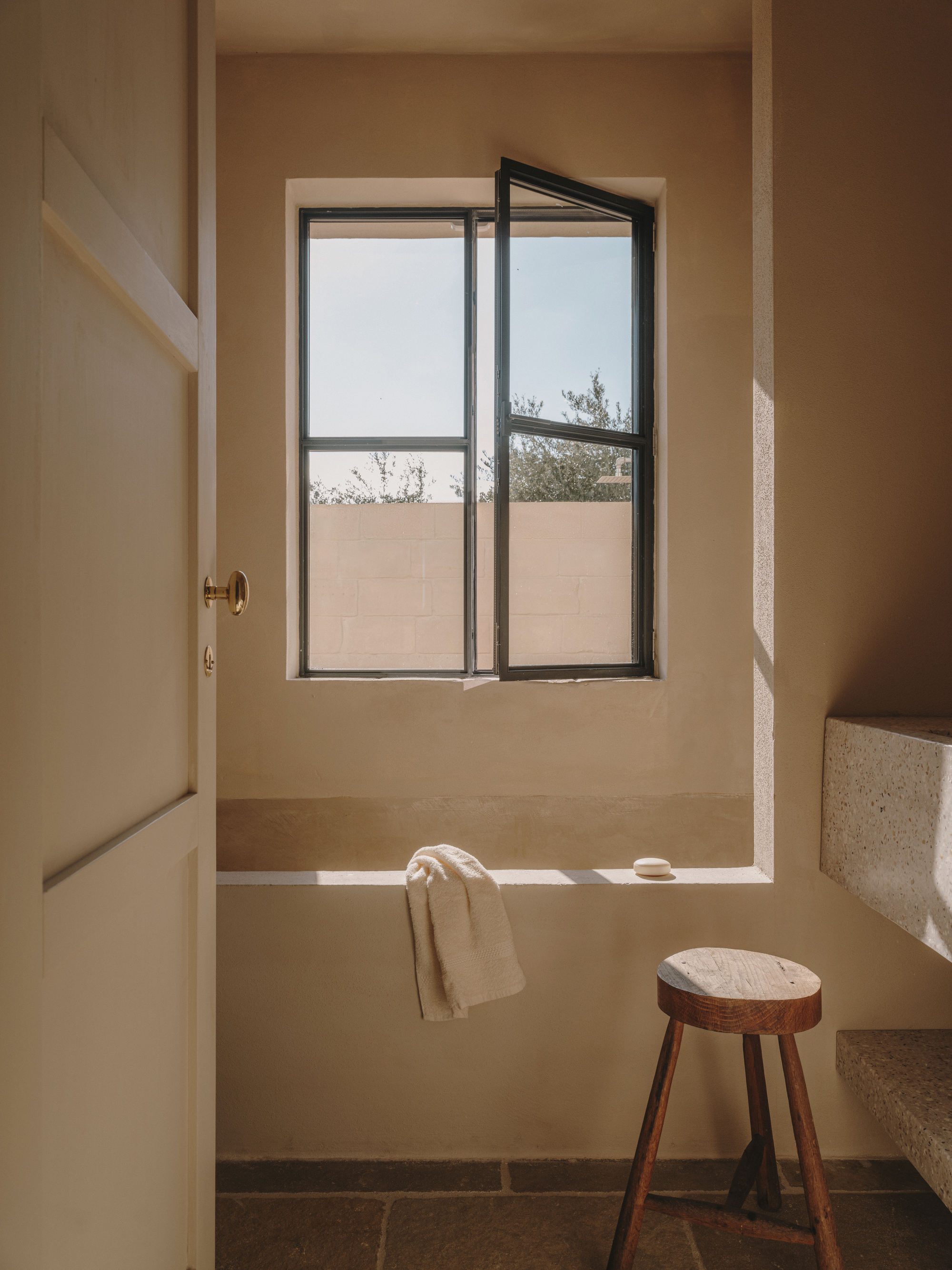 Built-in bathtub beneath a casement window overlooking a terrace with olive trees and wooden stool