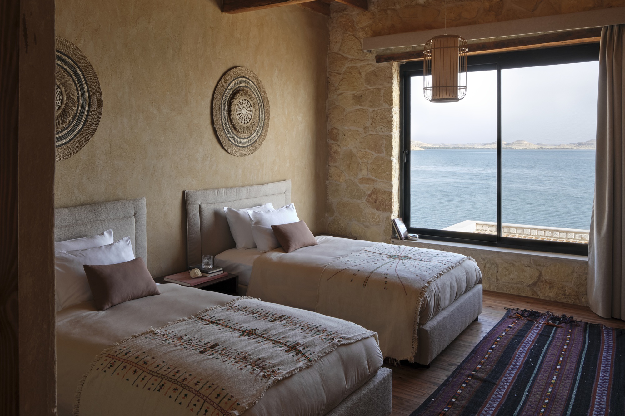 Twin beds with stone walls and exposed timber ceiling beams overlooking the water through large windows