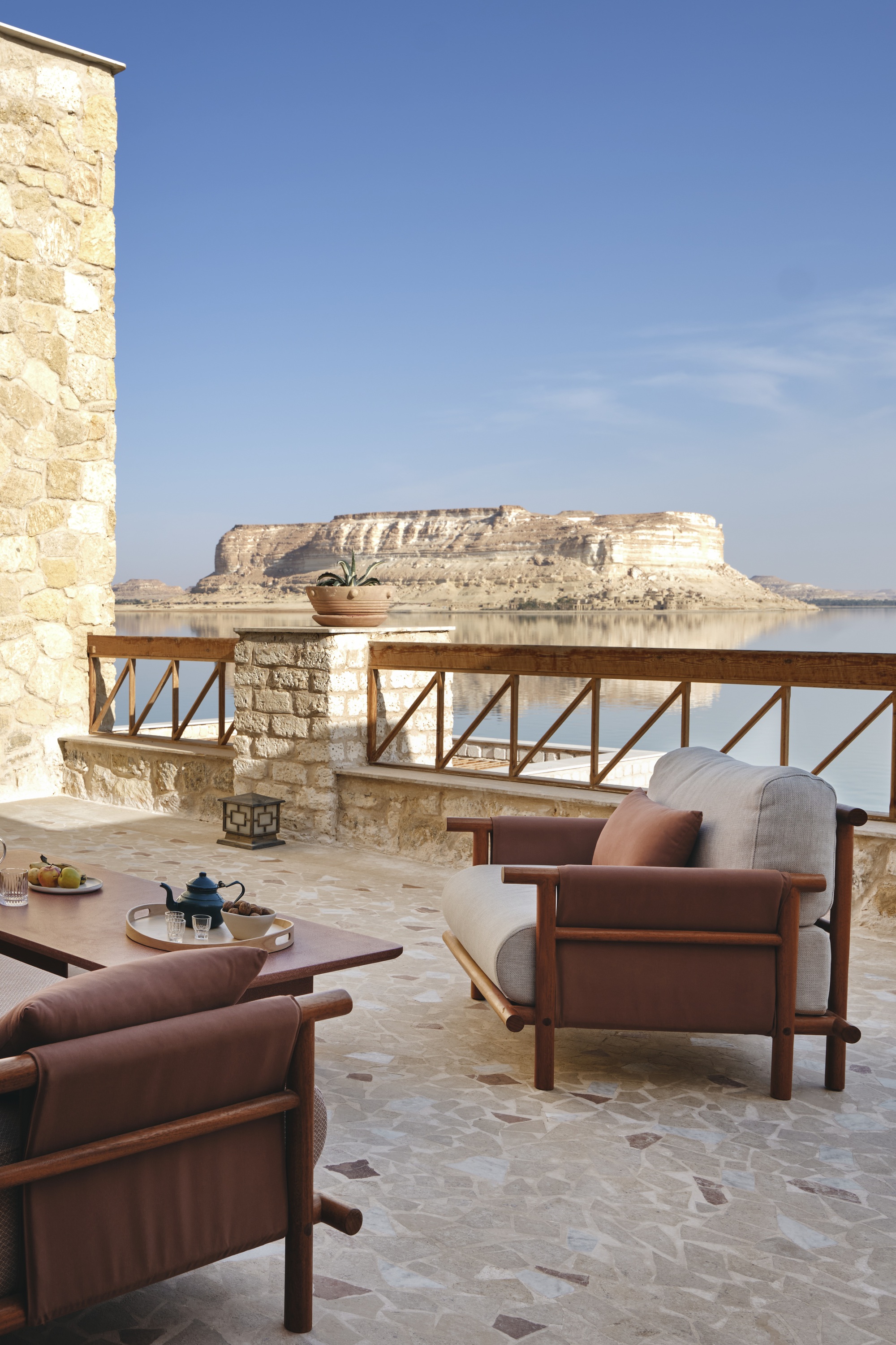 Limestone terrace with timber armchairs overlooking a distant mesa across calm water in bright daylight