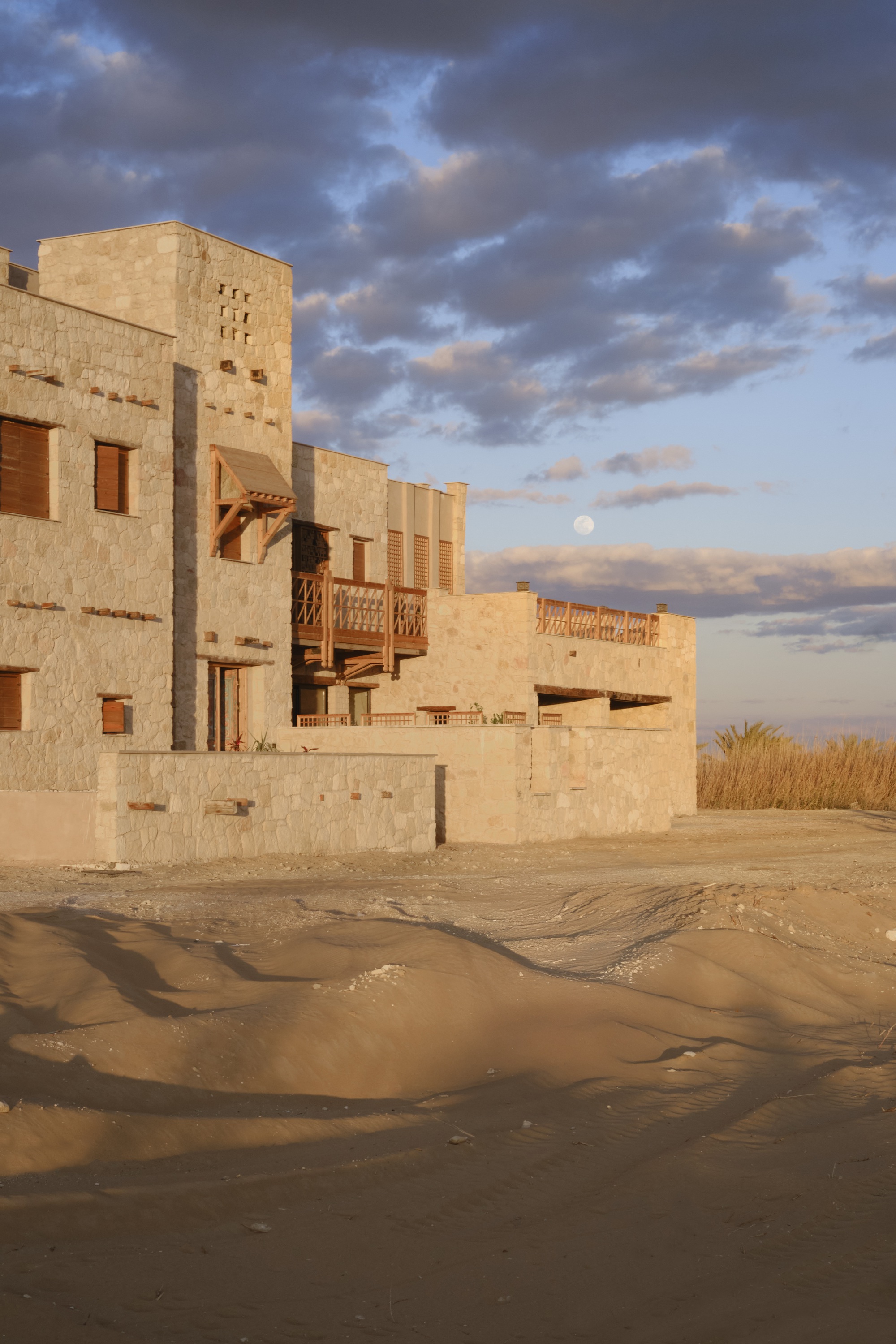 Clustered limestone volumes with timber balconies and openings rising from wet sand under evening clouds