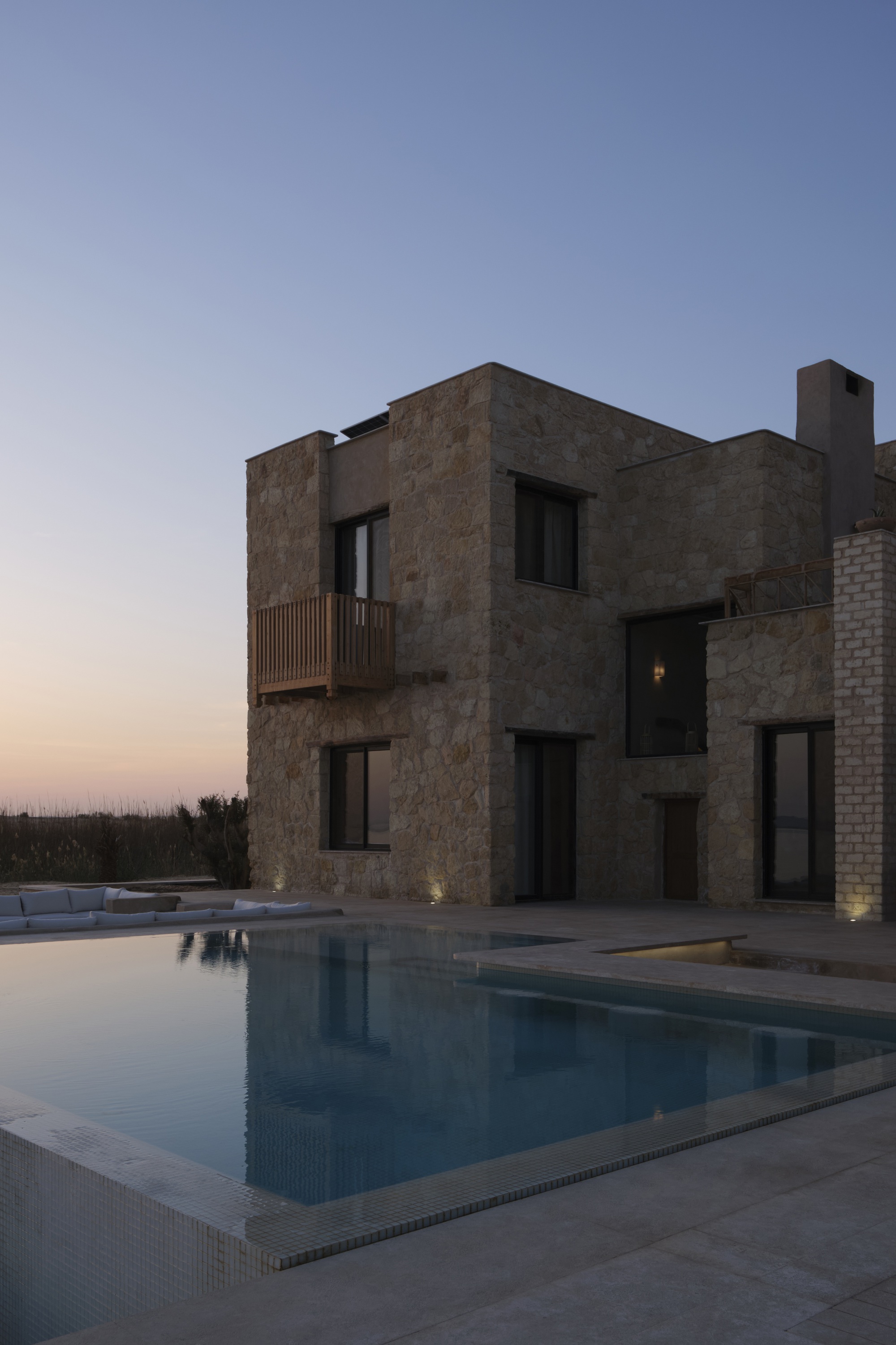 Stone facade with square openings and timber balcony beside a lit pool at twilight