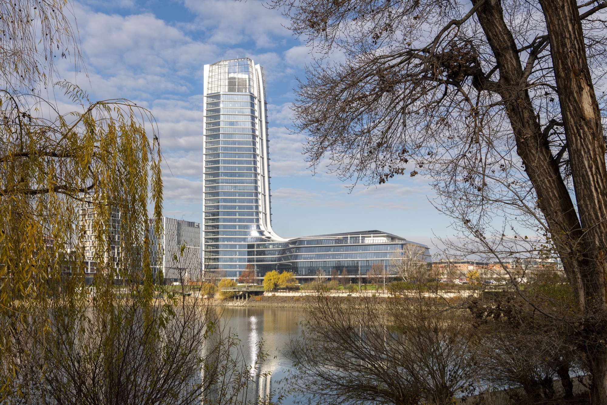 View across the river of the curved glass tower and low-rise base framed by willow and bare winter trees