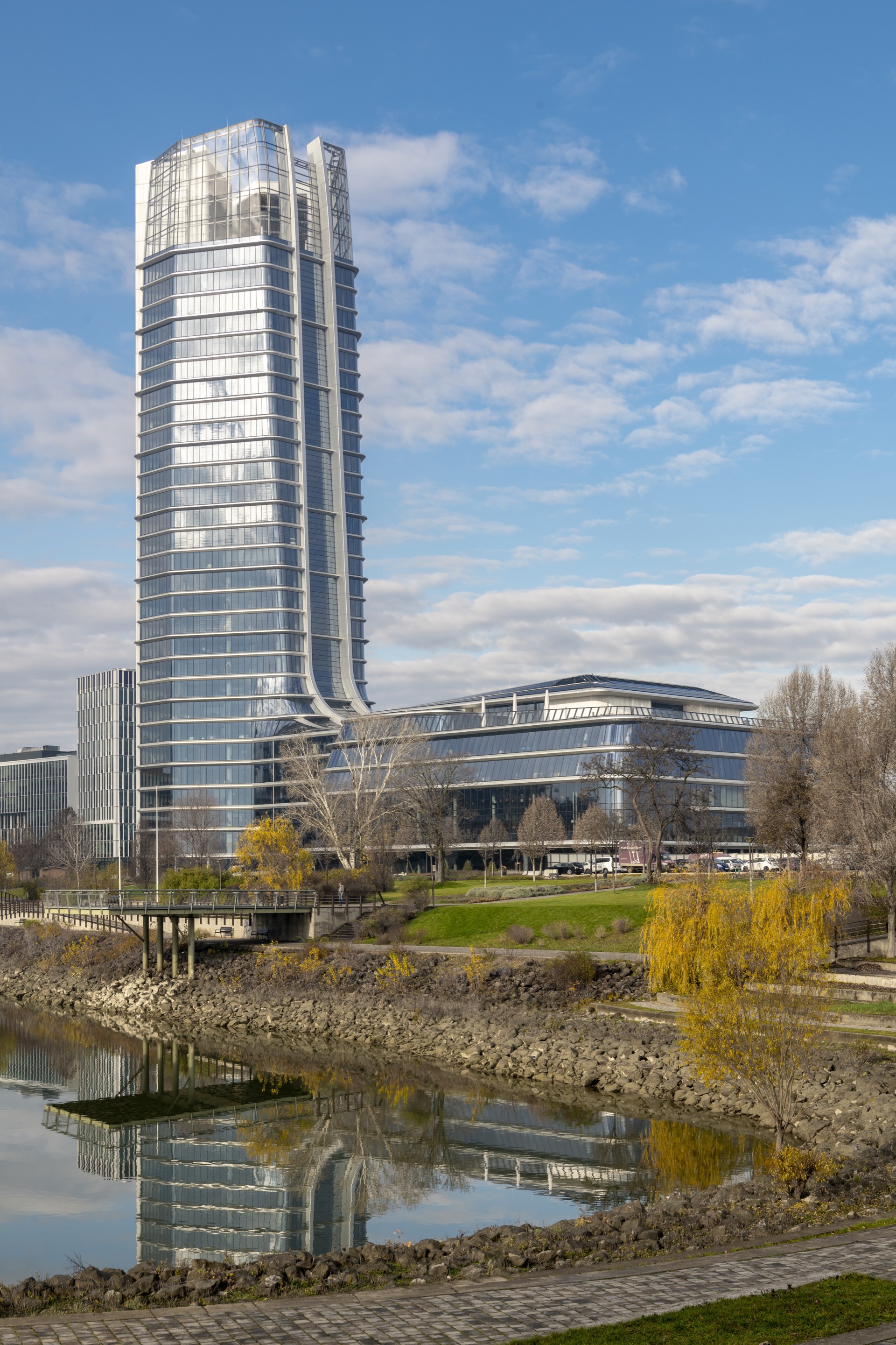 Waterfront view of the glass tower rising above the curved base with reflection in the foreground pond