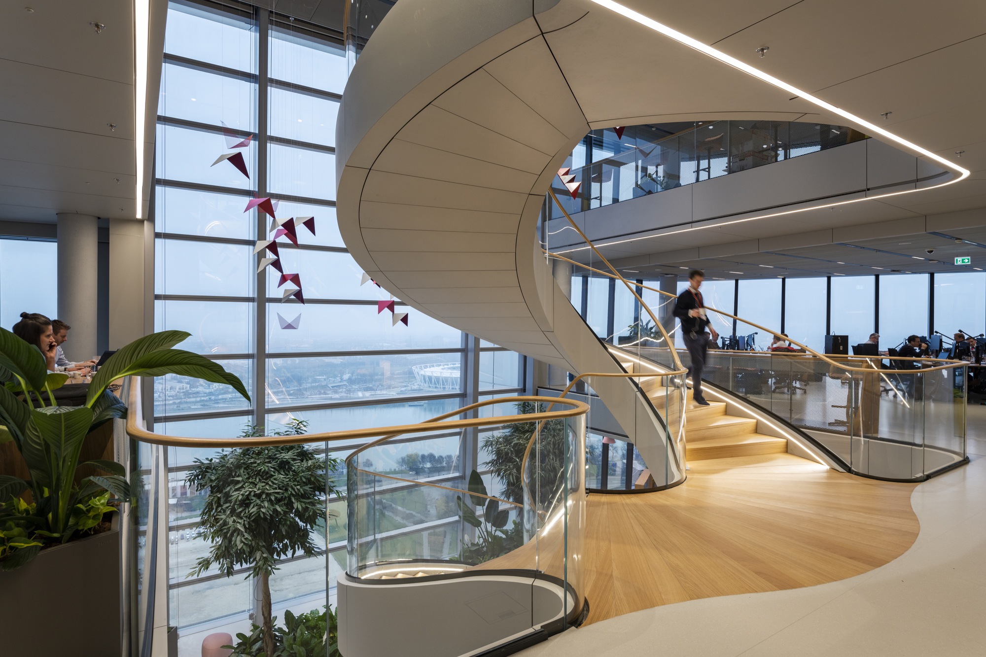 Spiral staircase with white paneled soffit and brass handrail ascending past floor-to-ceiling windows with red paper birds