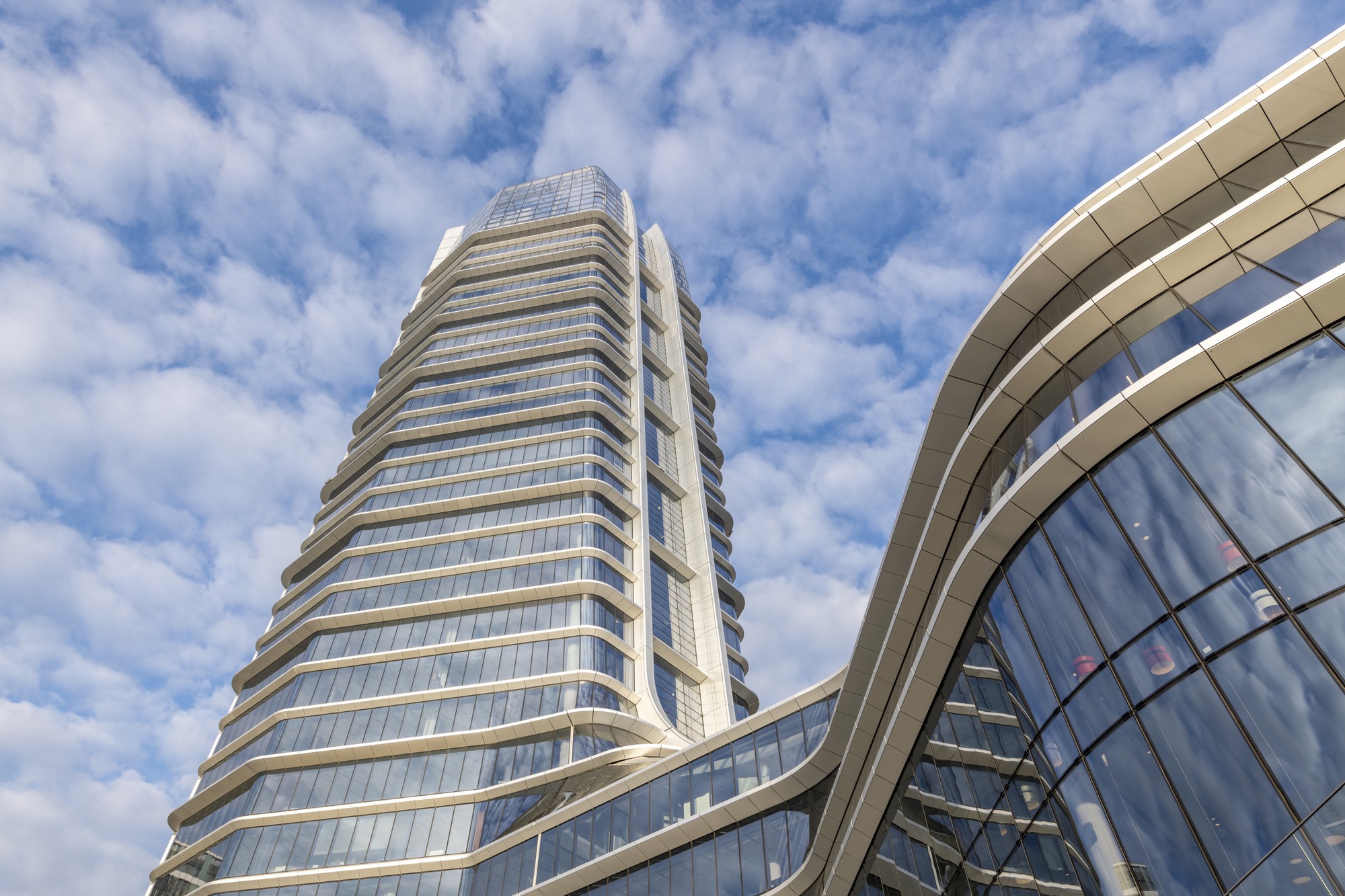 Upward view of the horizontally banded glass tower and curved podium facade under scattered clouds