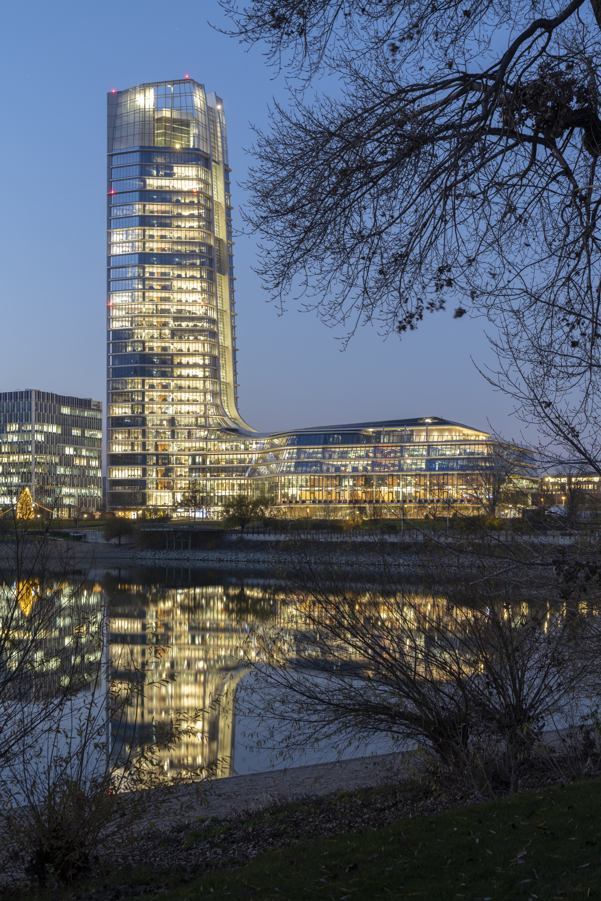 Evening view of the illuminated tower and waterfront podium reflected in the river framed by bare branches