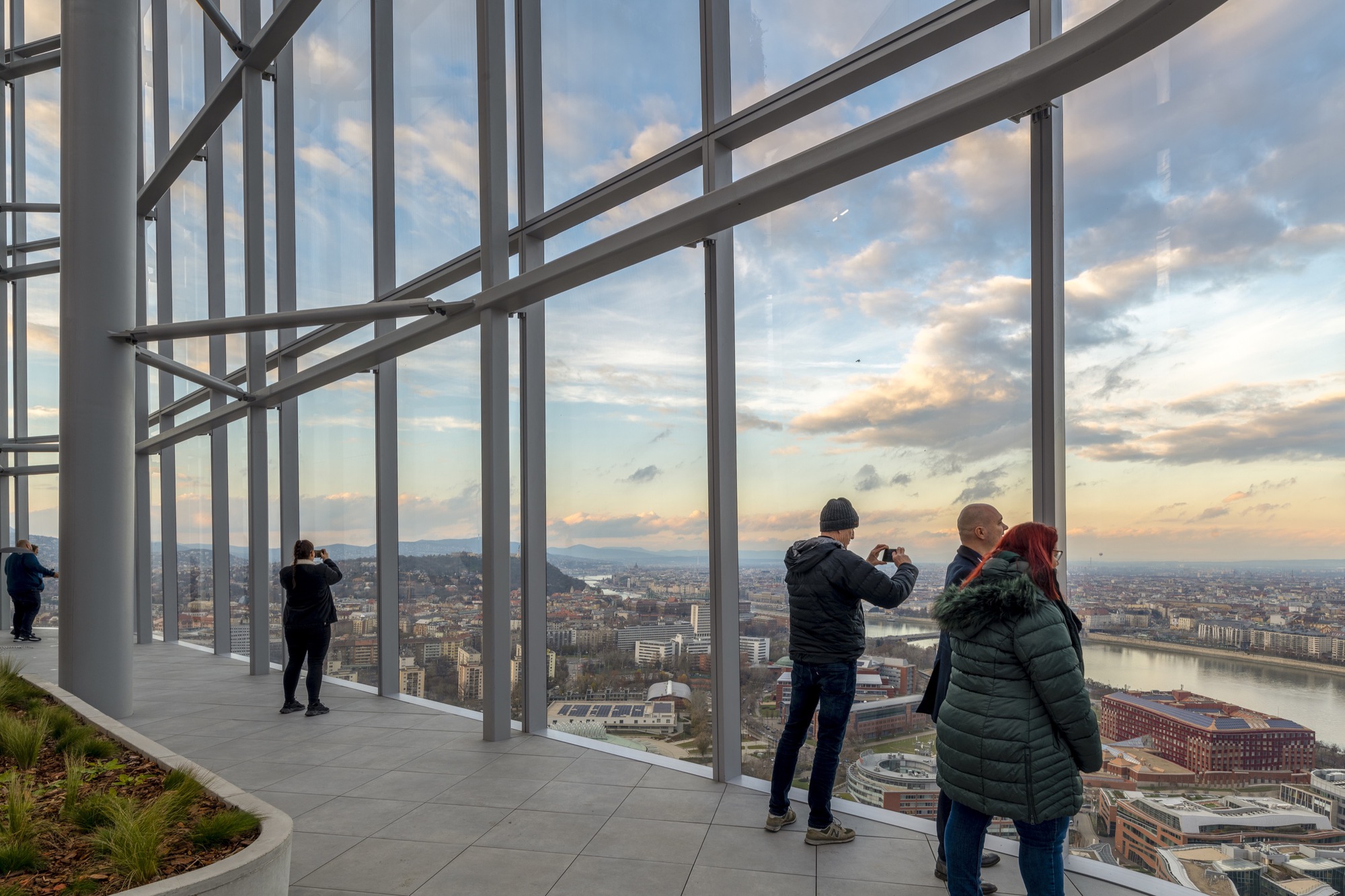 Floor-to-ceiling glazed observation deck with white steel bracing and visitors photographing the cityscape at sunset