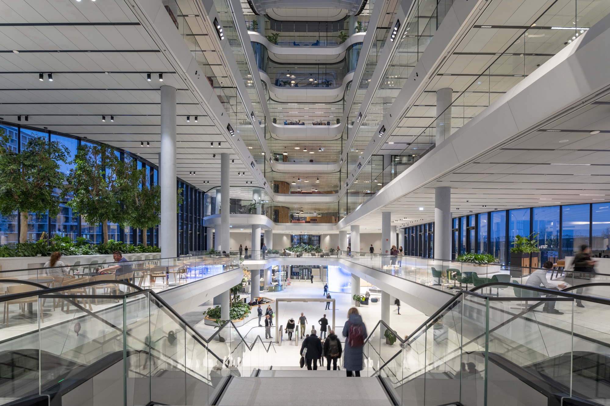 Multi-story atrium with glass-railed escalators descending toward a white marble lobby with planted trees