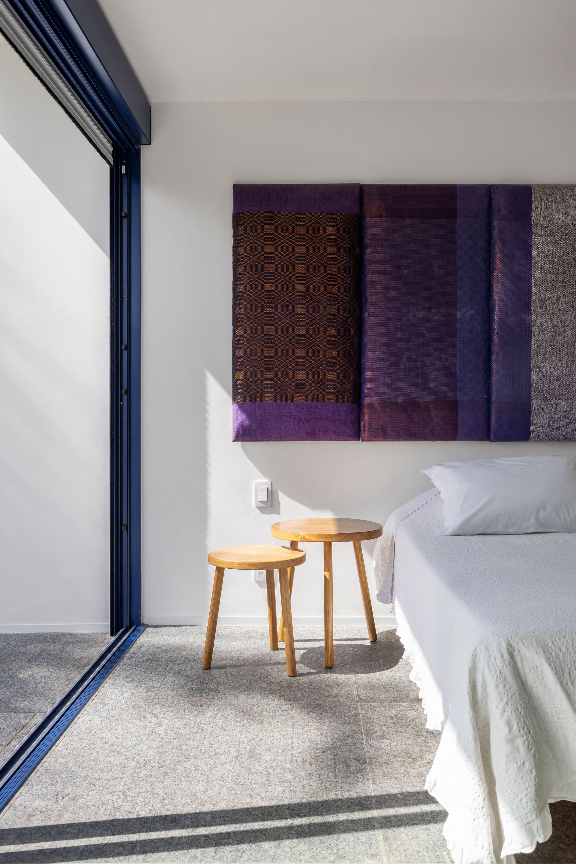 Bedroom corner with timber side tables and patterned textile artworks catching afternoon sunlight through dark-framed glazing
