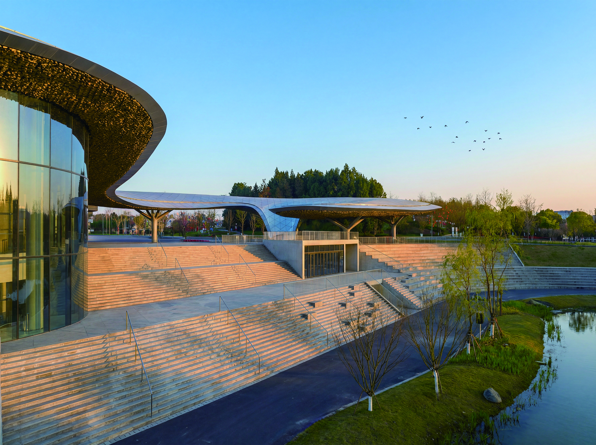 Timber plaza deck extending toward the water's edge at dusk with birds in flight