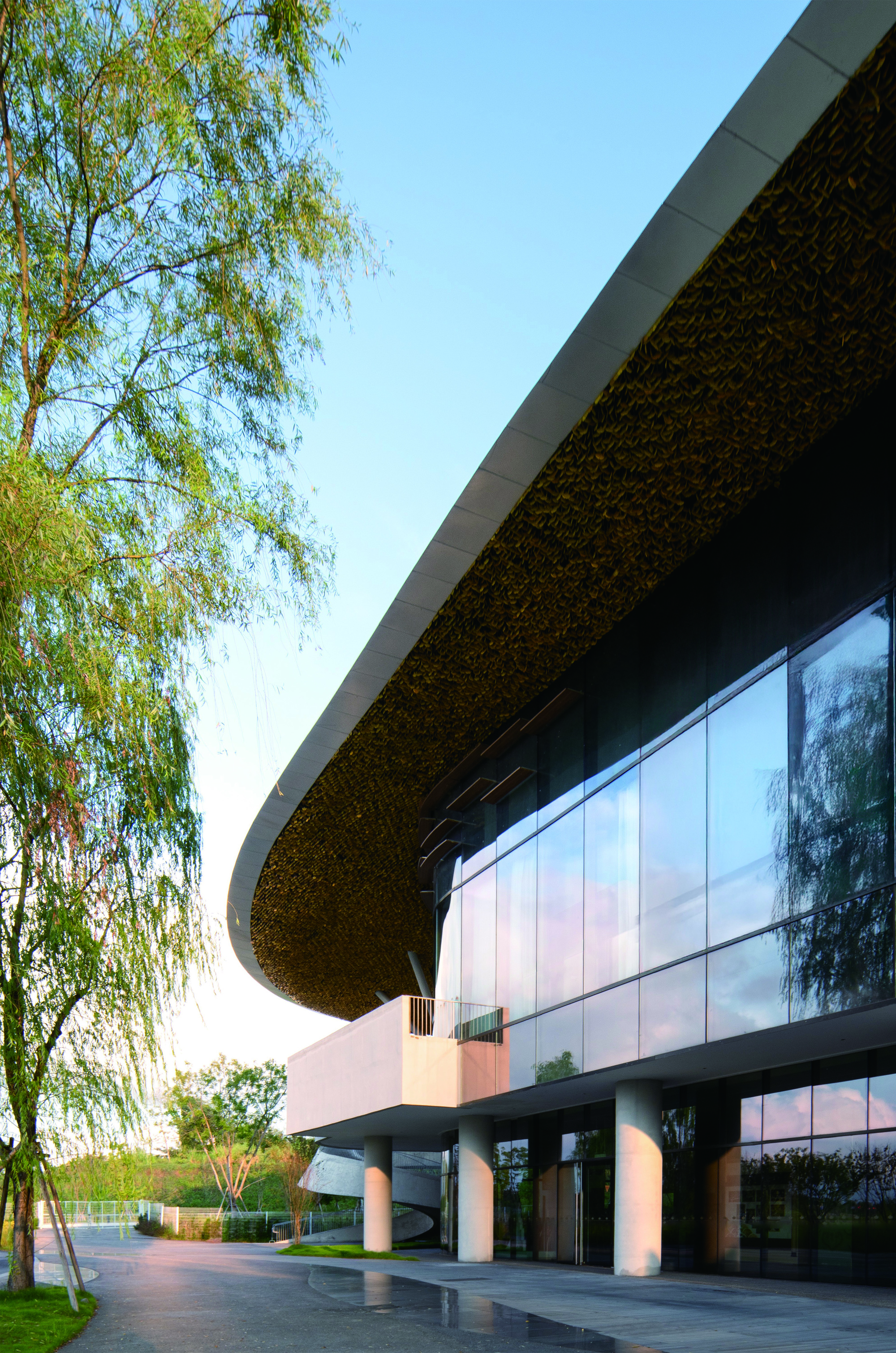 Glazed facade beneath a textured undulating roof canopy with white columns and willow trees at dusk