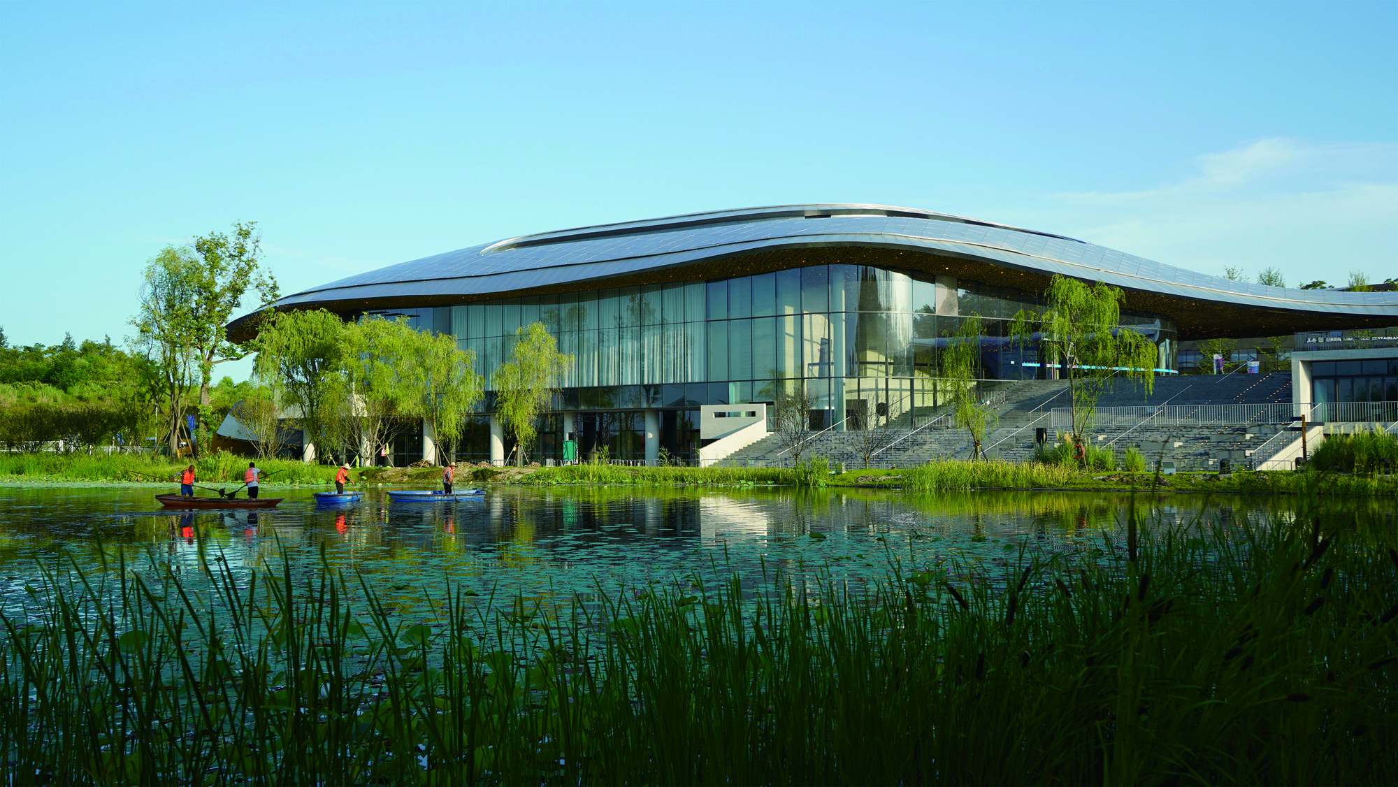 Glass curtain wall and curved roof reflected in a pond edged with tall grasses and visitors