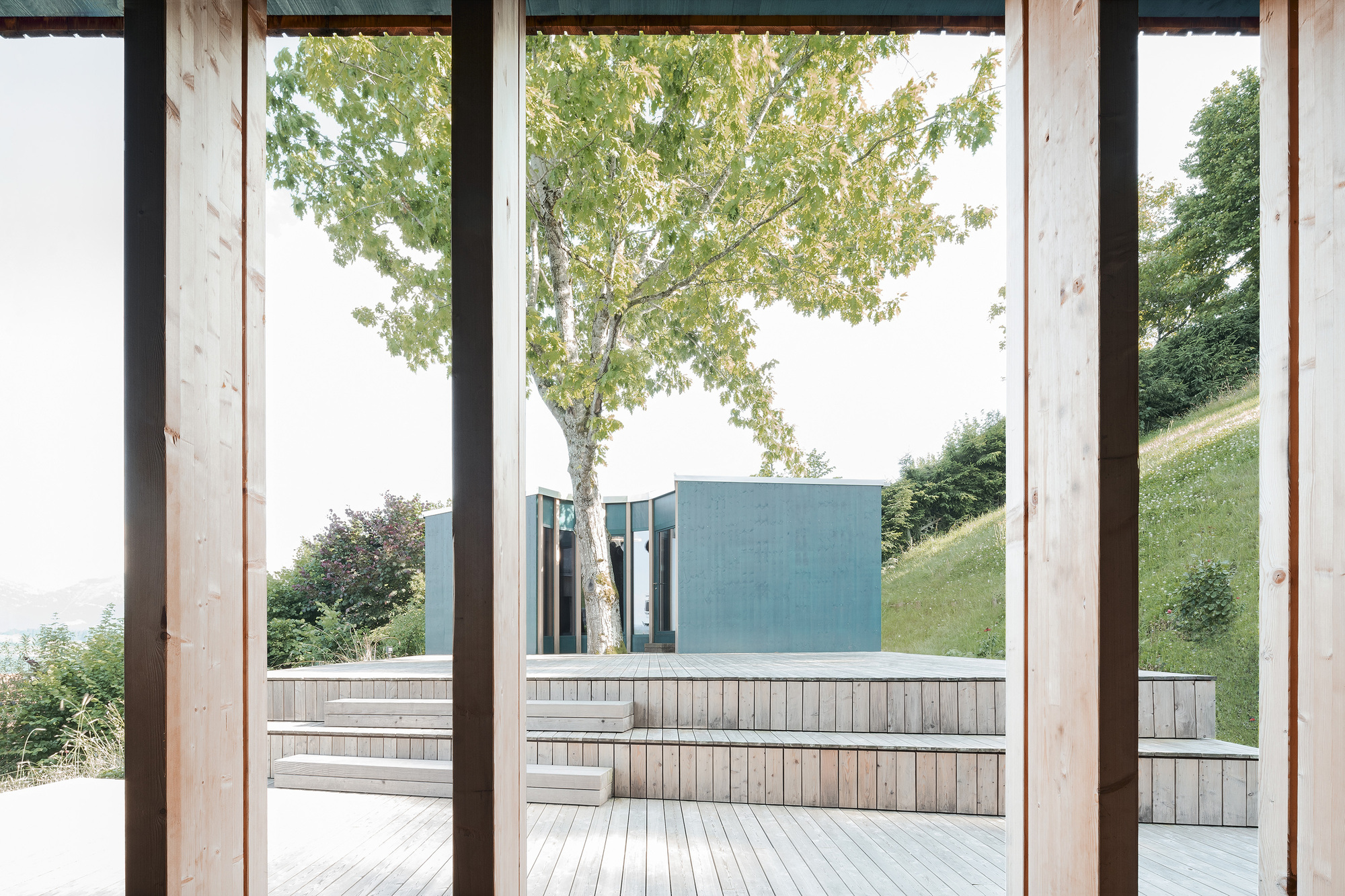 View from covered timber porch through black steel columns toward terraced deck and blue metal volume