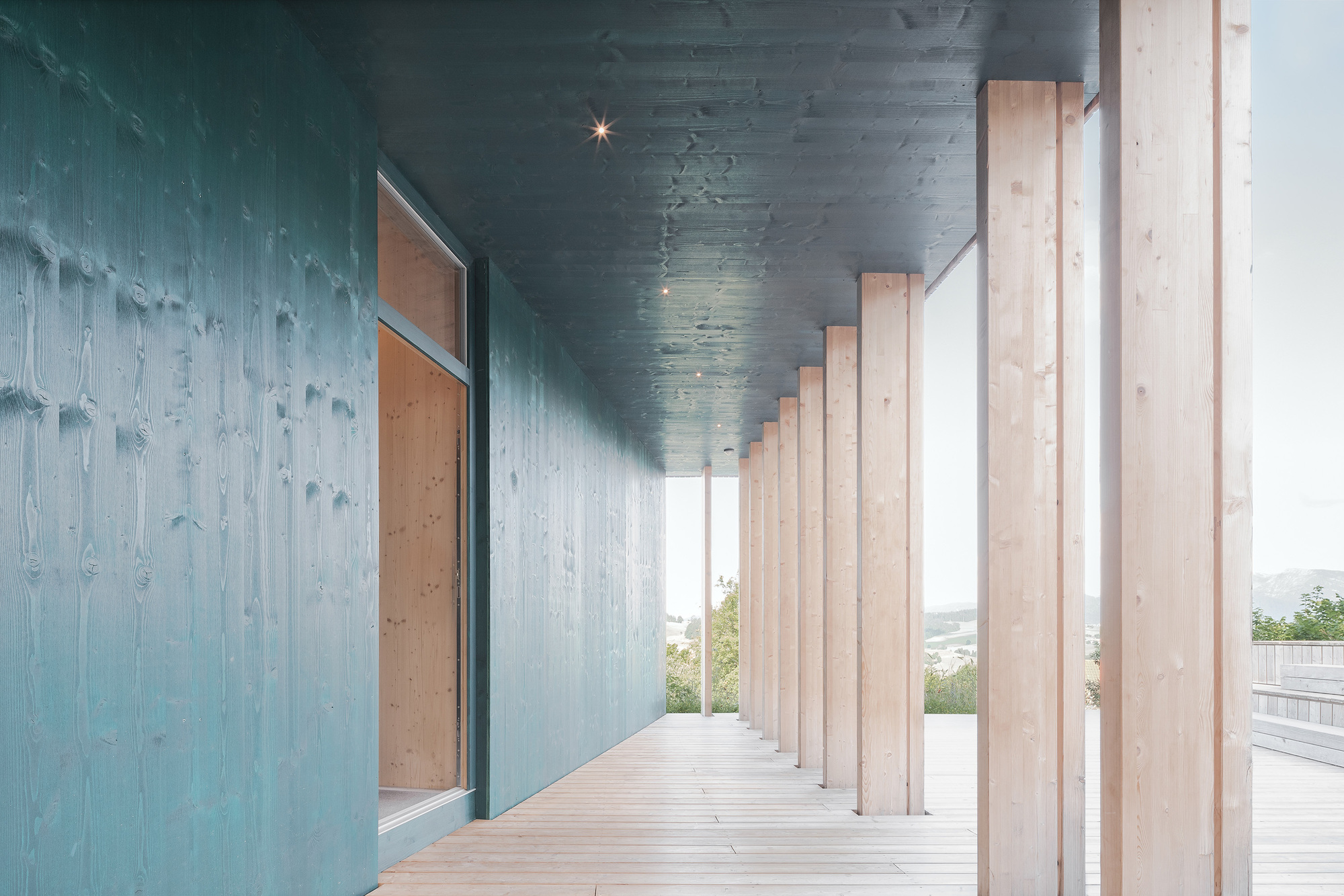 Covered colonnade with timber columns between blue stained wood walls and decking under grey sky
