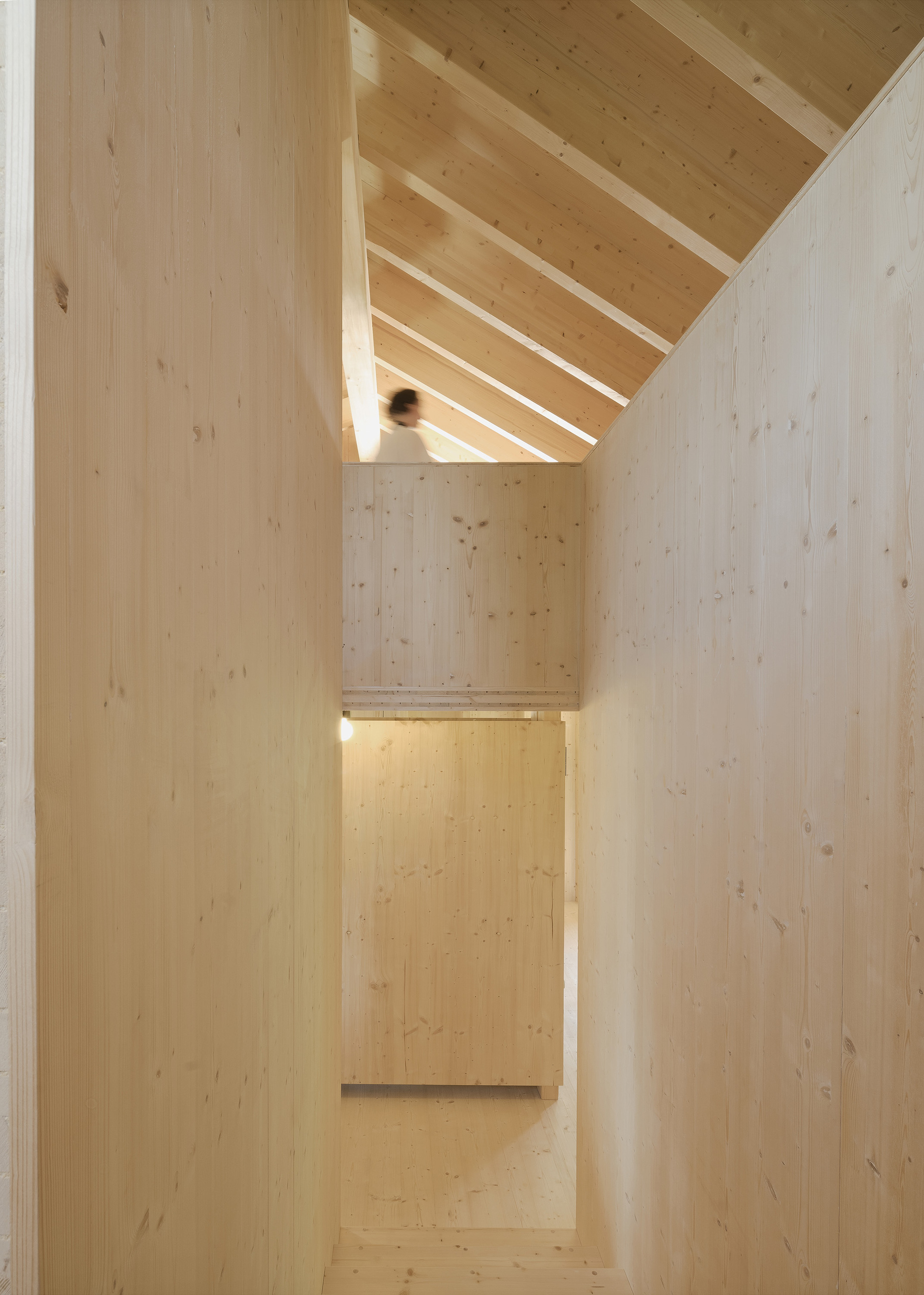 Light wood stairwell with exposed beams and a figure ascending beneath the pitched roof