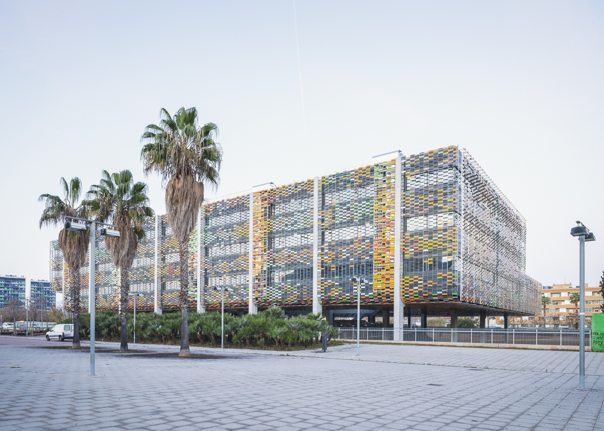 Perforated facade with multicoloured ceramic panels on white columns viewed from a paved plaza with palm trees