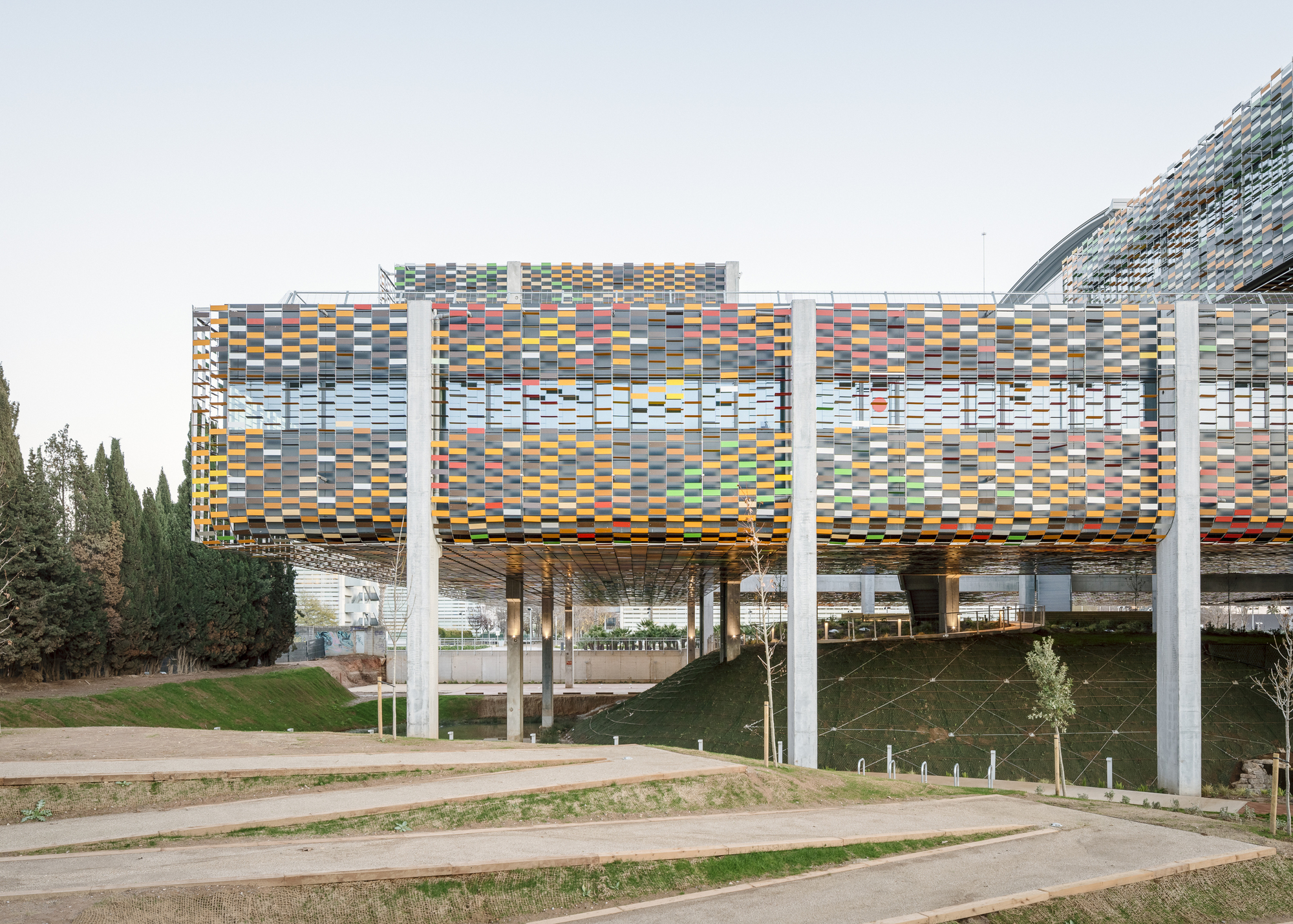 Elevated structure with multicoloured ceramic screen cladding supported by white concrete columns above a landscaped slope
