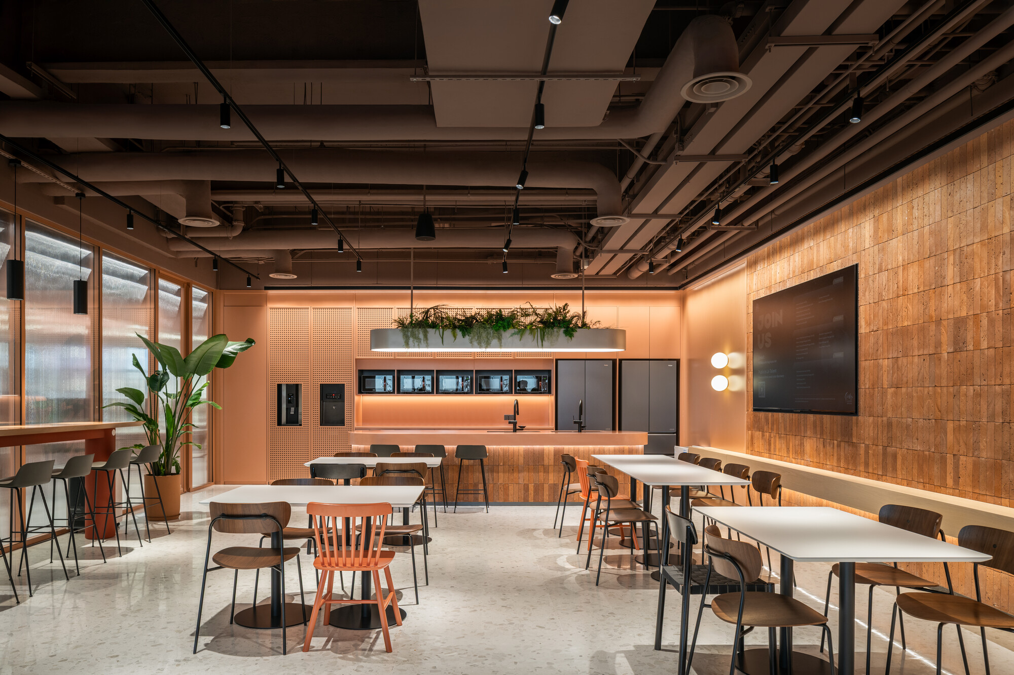 Communal dining area with scattered tables and an orange-lit kitchenette beneath an exposed structural ceiling