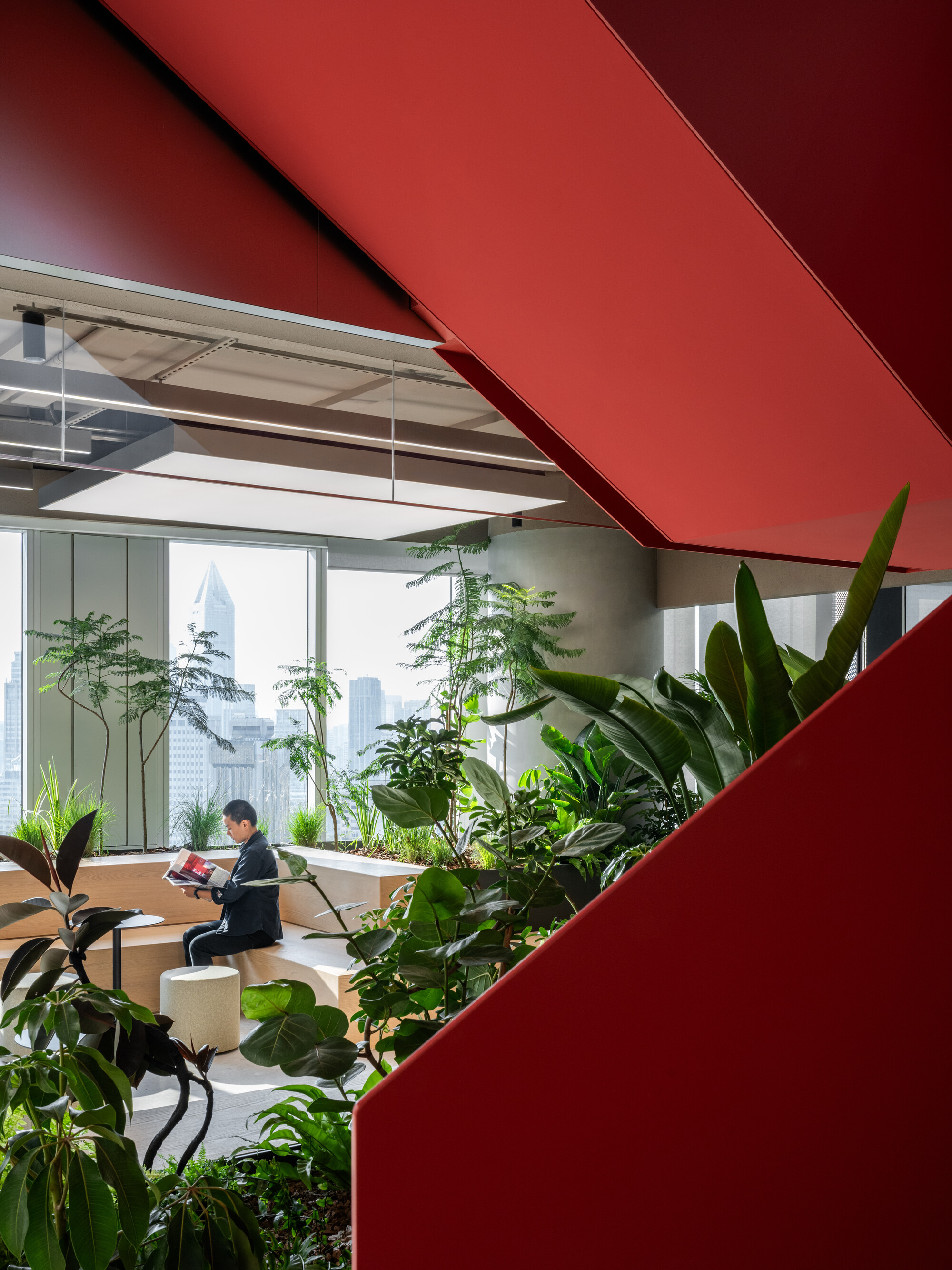 Interior workspace framed by red sculptural fins with planted beds and a person seated near floor-to-ceiling windows
