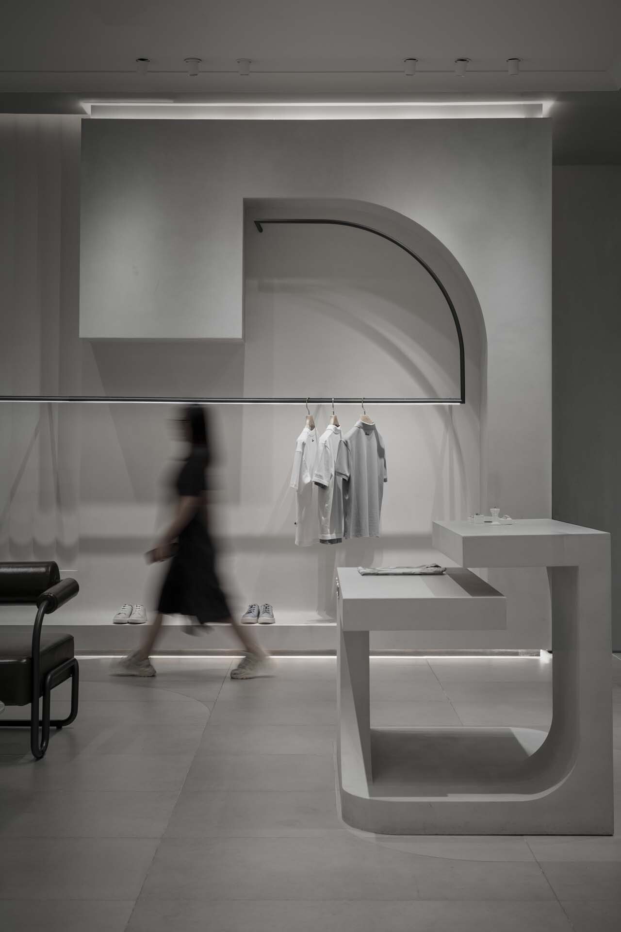 Arched display niche with clothing rack and blurred figure walking past white cantilevered platform