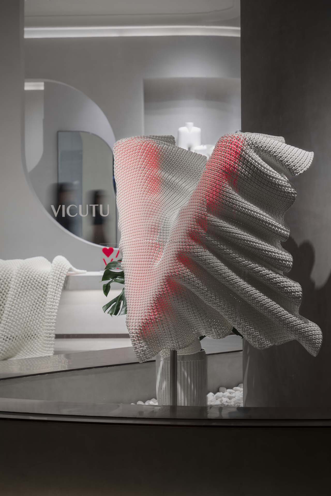 Close-up of red and grey mesh fabric sculpture on glass table beside circular mirror
