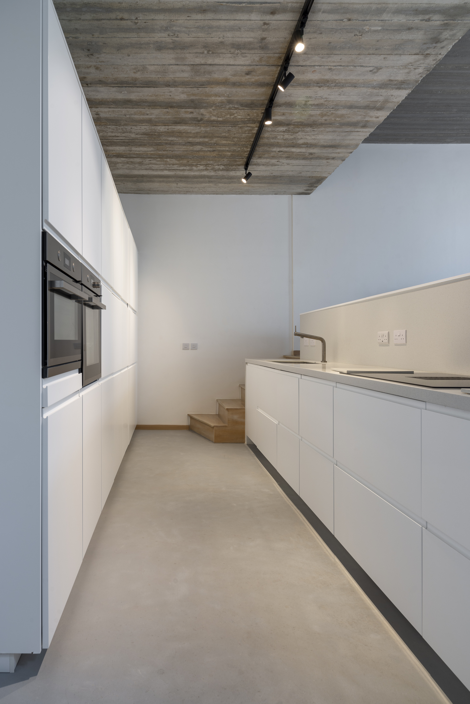 Galley kitchen with white cabinetry and timber steps beneath board-formed concrete ceiling and black track lighting