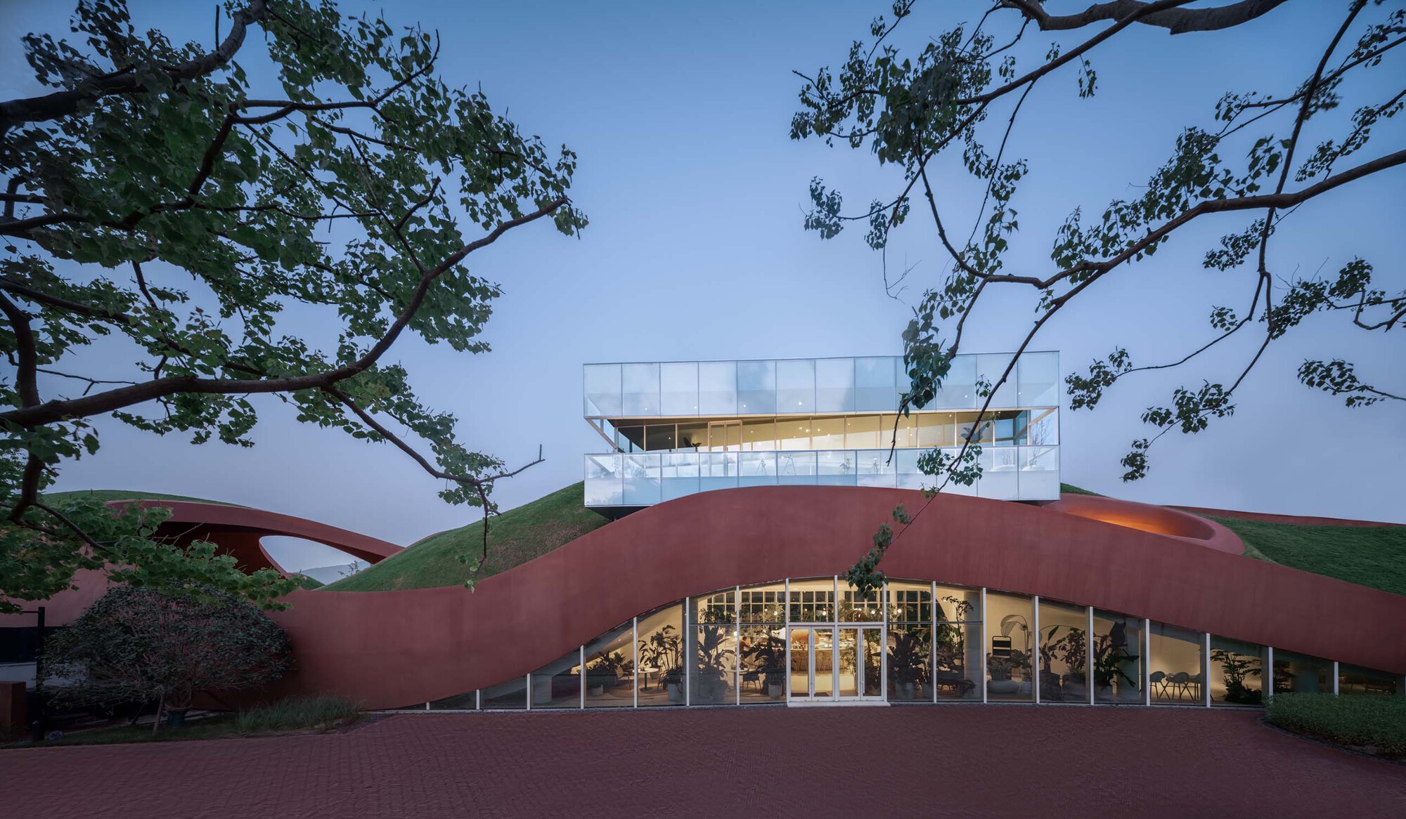 Main entrance with glazed facade beneath the sweeping roof canopy framed by mature trees at dusk
