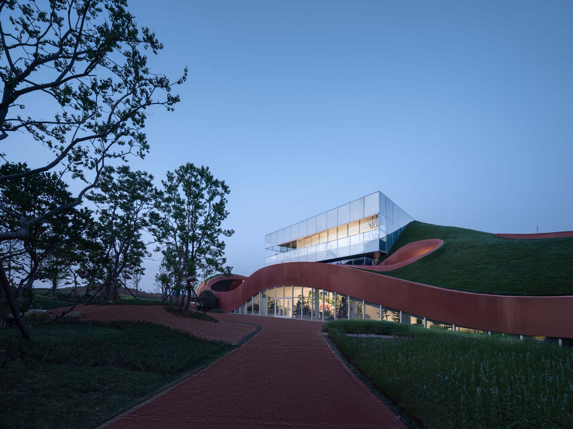 Approach path winding through the undulating roof forms toward the glass pavilion at twilight
