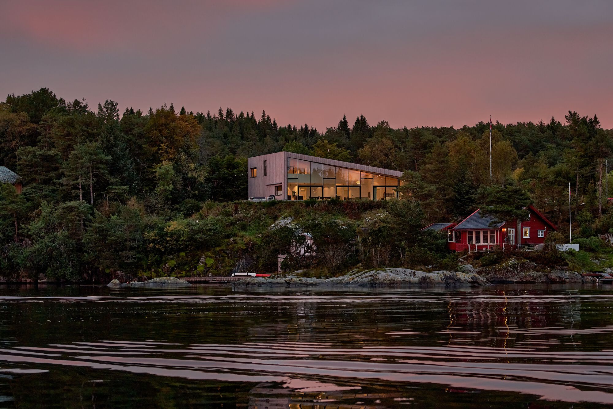 Waterfront view of the illuminated glass facade perched above rocky shoreline at dusk