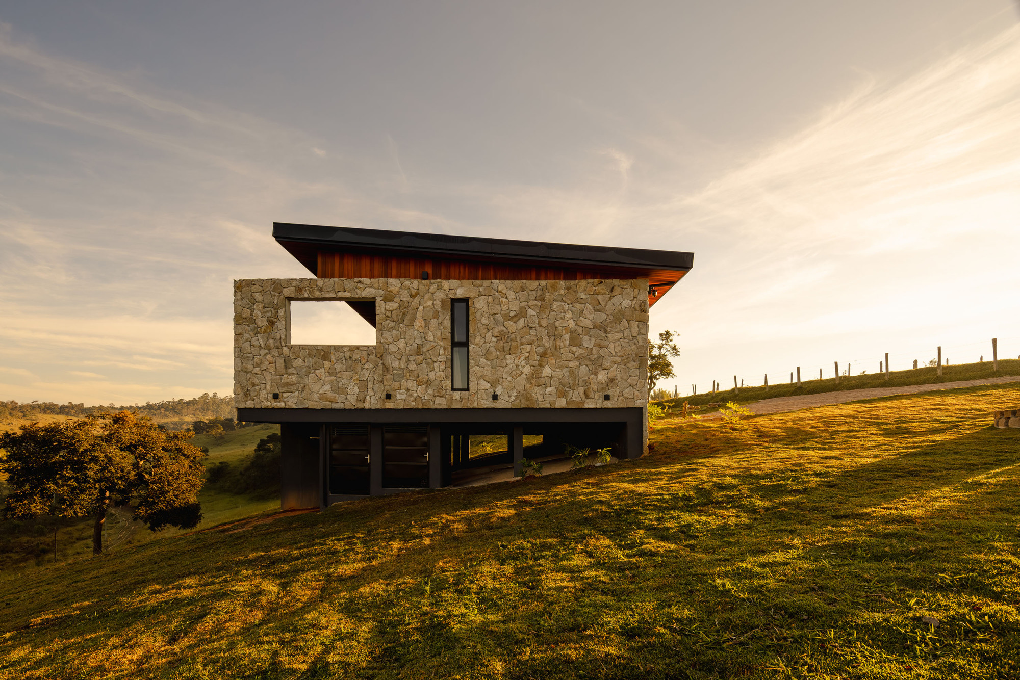 Two-story volume with stacked stone upper level and recessed glazed base on a grassy hillside at dusk