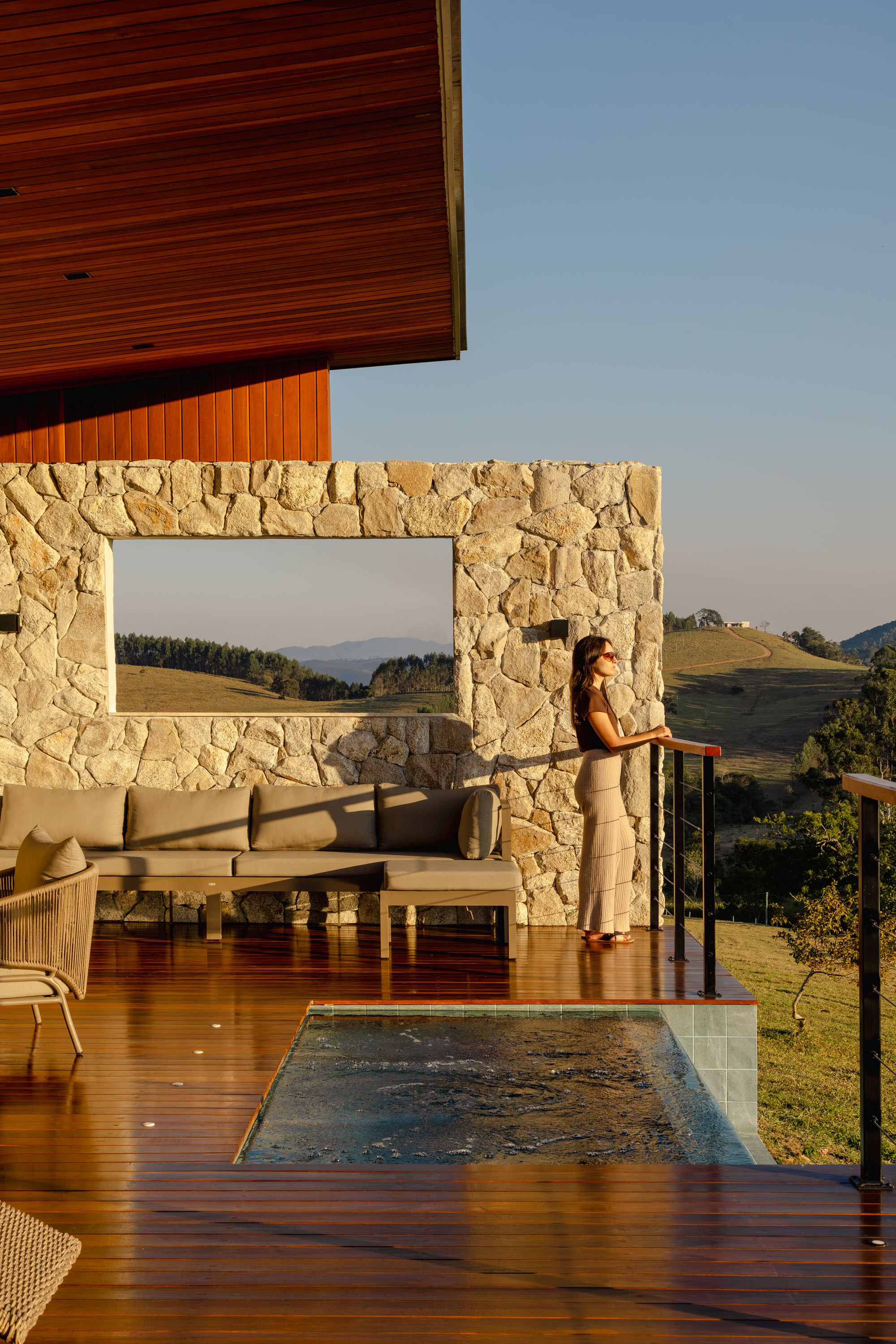 Covered timber deck with stone-framed opening and a person standing by the glass railing at sunset