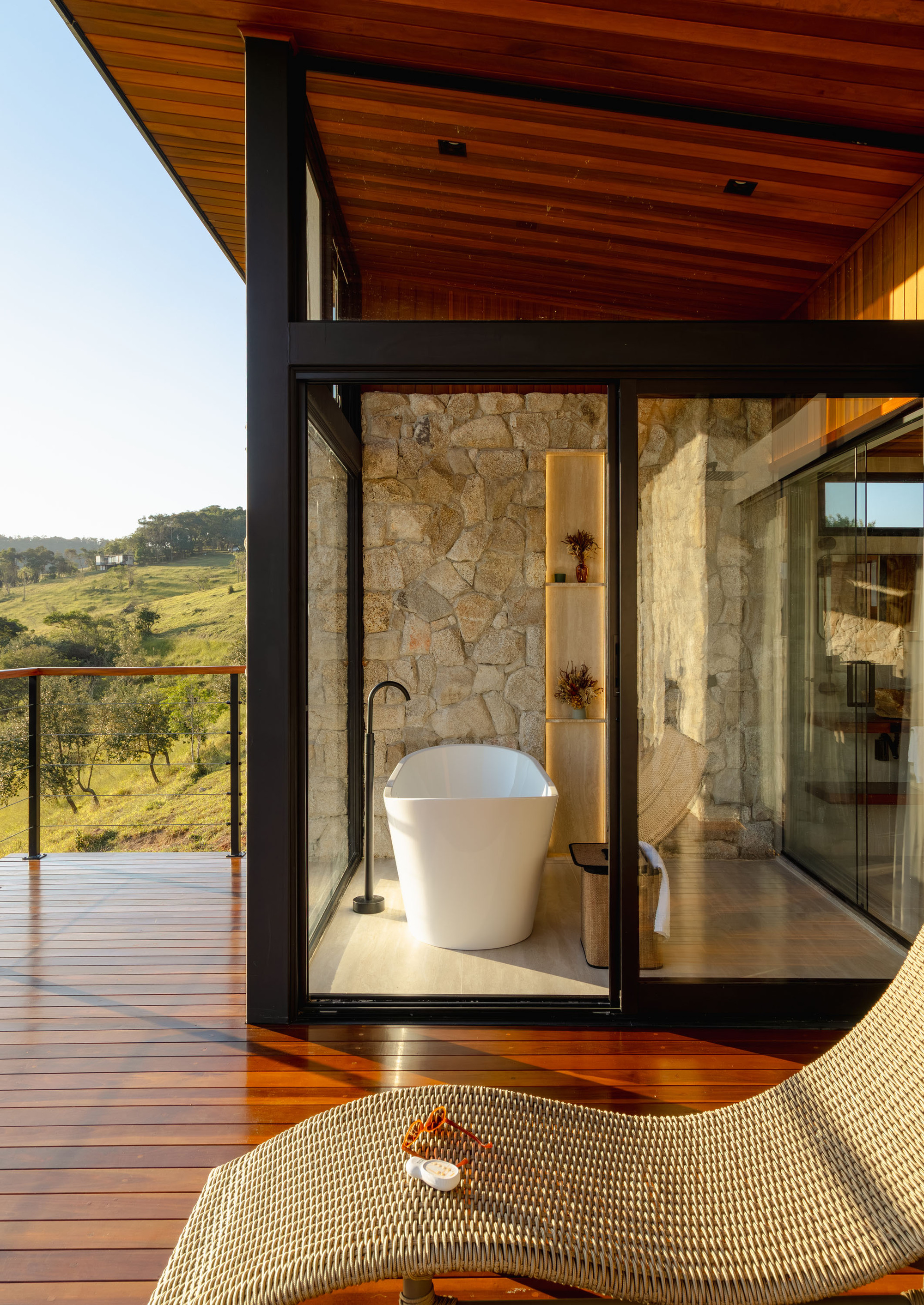 Glass-enclosed bathtub alcove with stone walls opening to timber deck and valley views at sunset