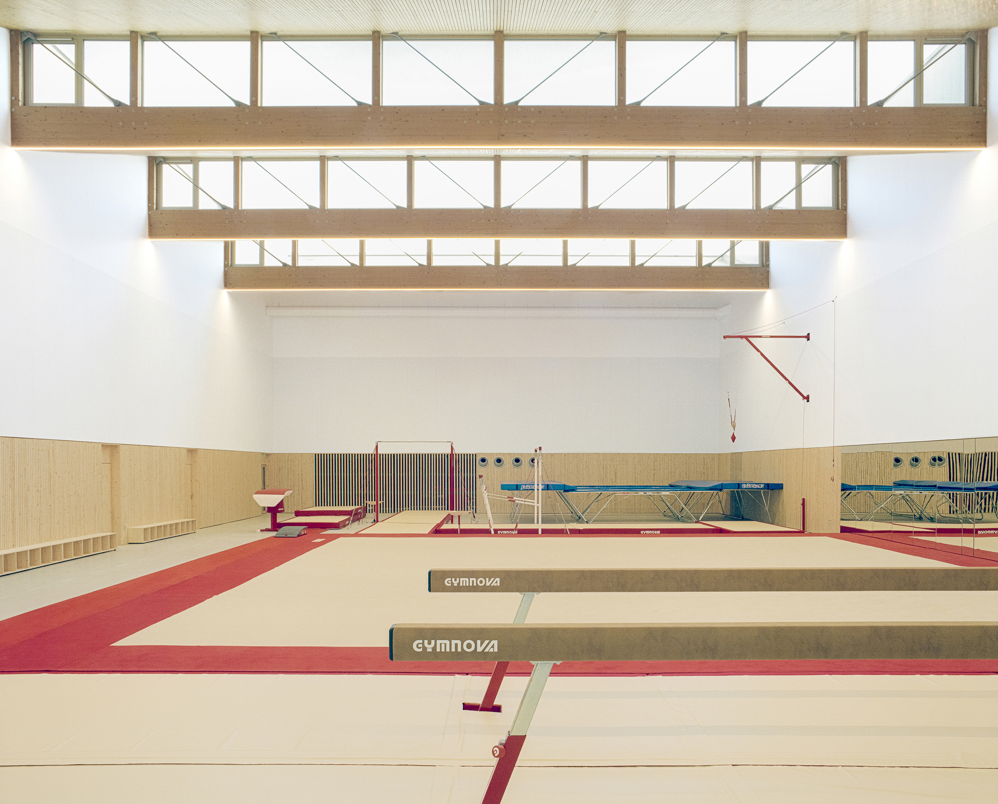 Gymnastics hall with tiered timber clerestory monitors above beams and maroon floor mats