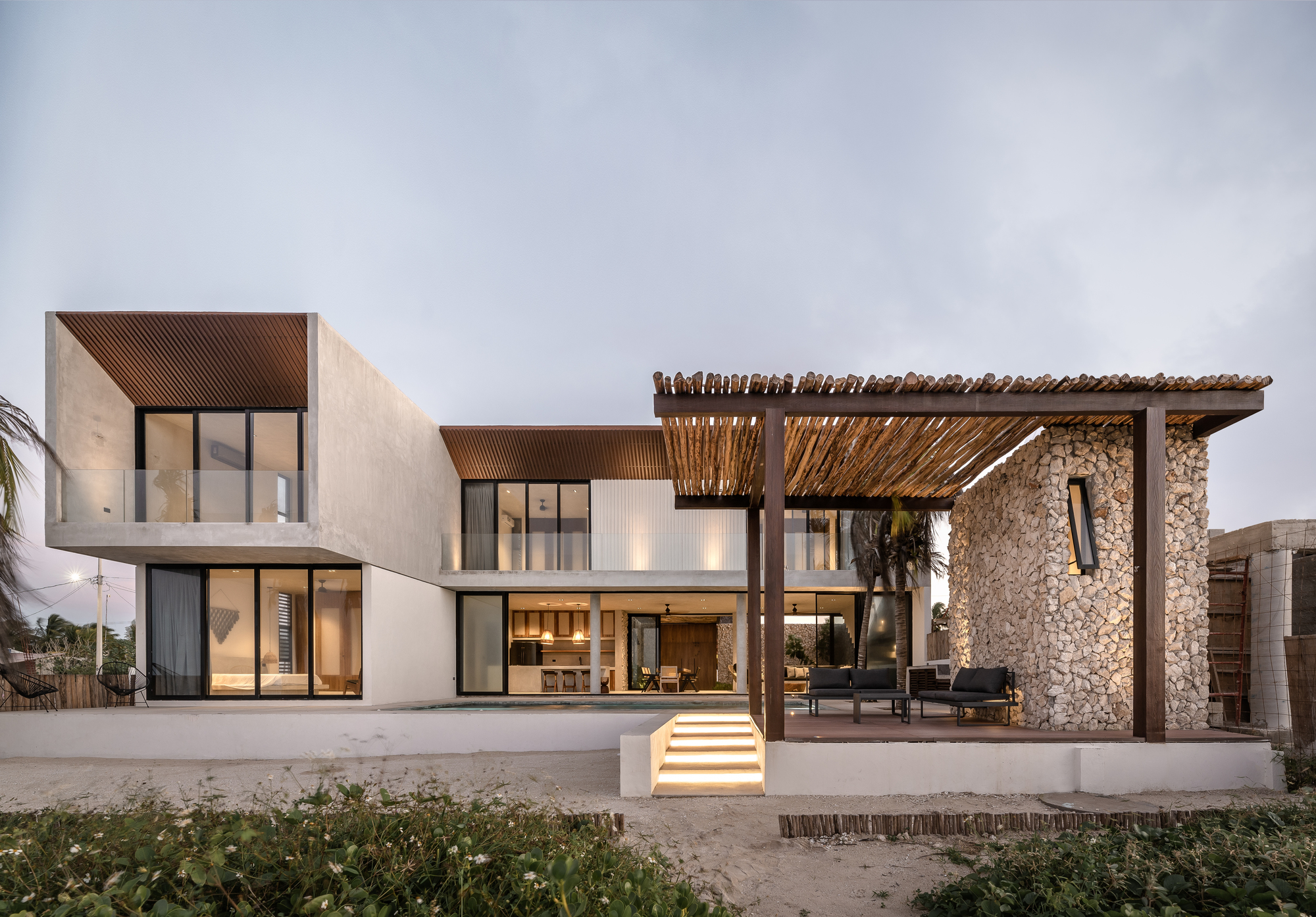 Rear facade showing white rendered volumes with a gabion wall pergola and illuminated entry steps at dusk