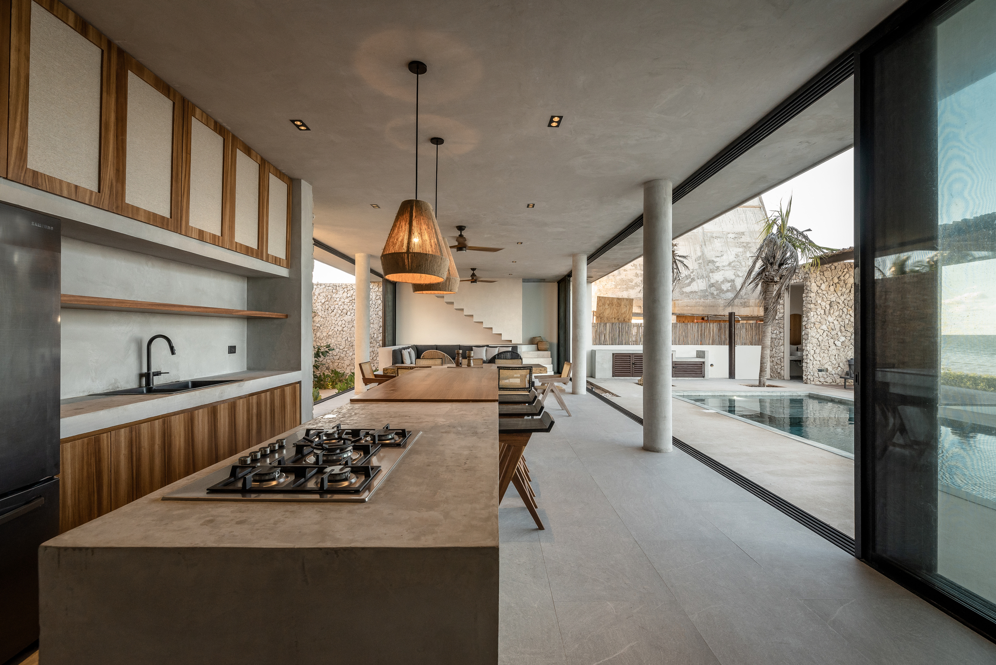 Open kitchen with concrete island and floor-to-ceiling glazing overlooking a pool and gabion walls