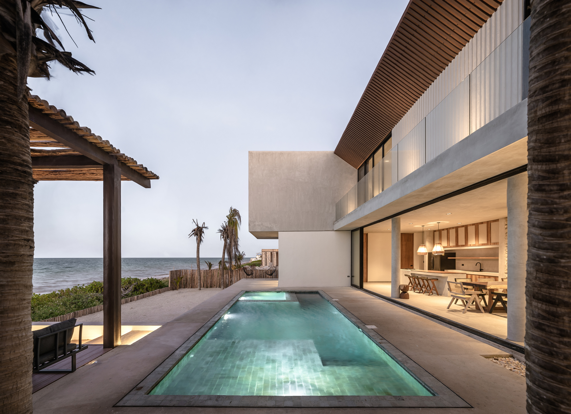 Pool deck with ocean view flanked by thatched shelter and cantilevered volume at dusk