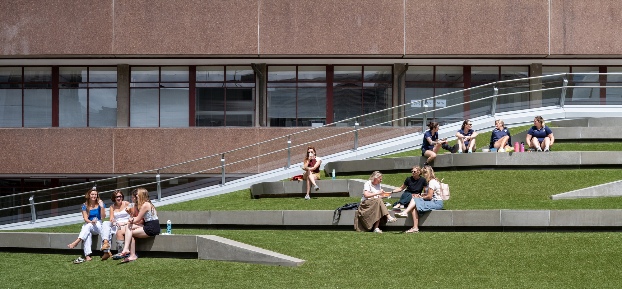 Tiered outdoor seating with alternating grass and concrete steps occupied by students in sunshine