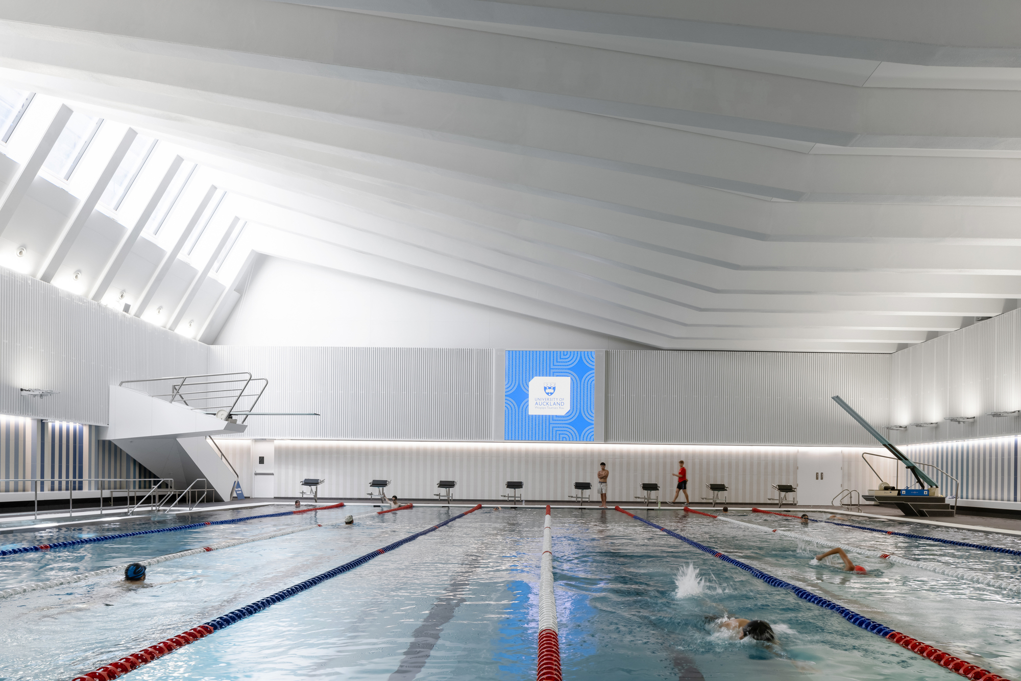 Indoor lap pool with white ribbed ceiling and swimmers in lanes under overhead skylights