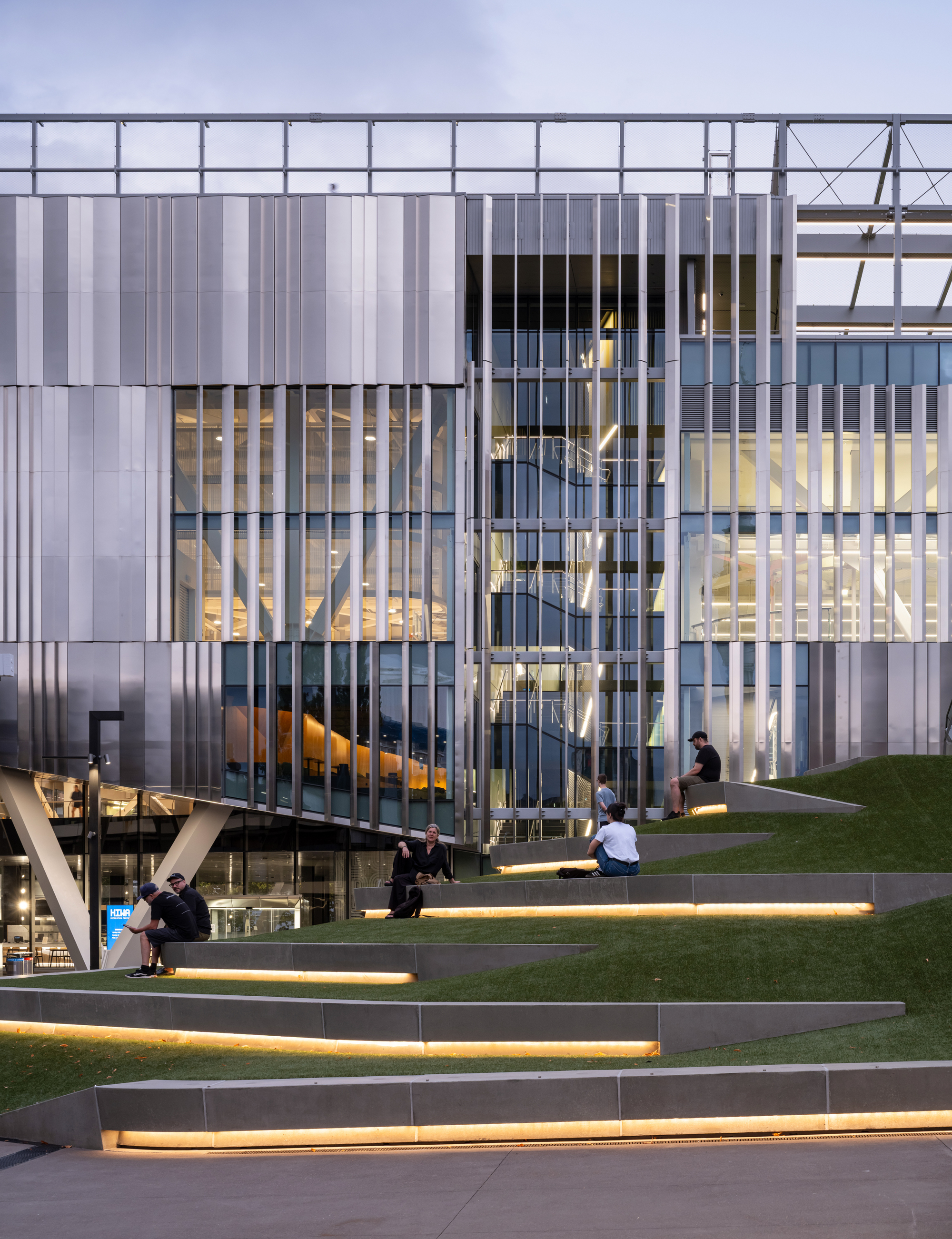 Exterior view of translucent facade with vertical fins and stepped lawn with visitors at dusk